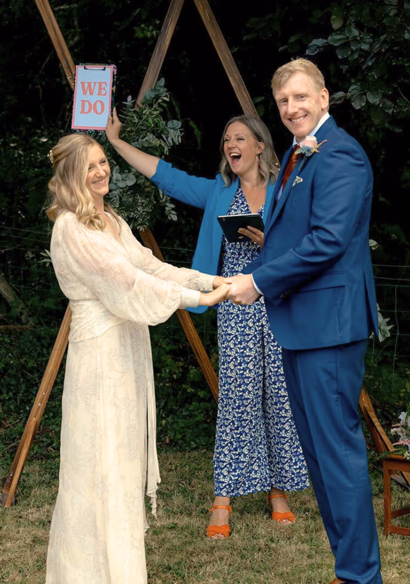 Bride Katie and groom Seb holding hands during their North Wales outdoor wedding ceremony, with celebrant Kate Rostance leading the vows and holding up a playful “We Do” sign.