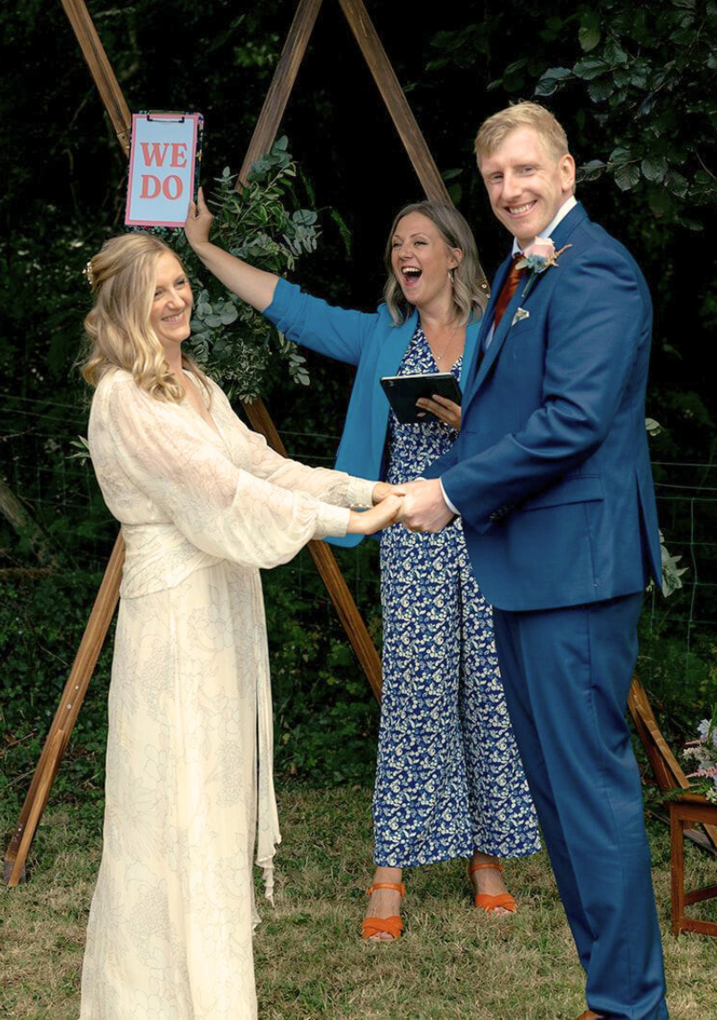 Bride Katie and groom Seb holding hands during their North Wales outdoor wedding ceremony, with celebrant Kate Rostance leading the vows and holding up a playful “We Do” sign.