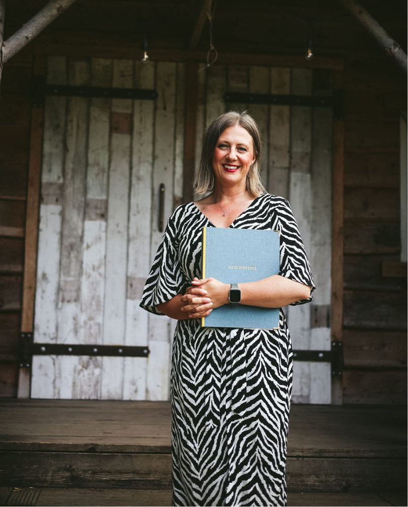Wedding celebrant Kate Rostance standing in front of rustic barn doors, holding her ceremony folder and smiling, ready to lead a personal and modern wedding ceremony.