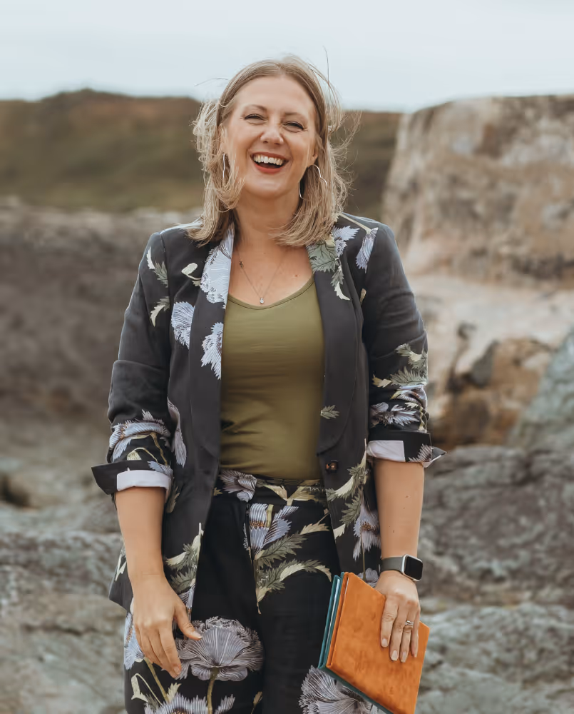 Kate Rostance, a wedding and funeral celebrant, smiling warmly outdoors against a rocky backdrop. She is wearing a floral patterned suit with a green top, holding a leather-bound notebook, and looks relaxed and approachable.