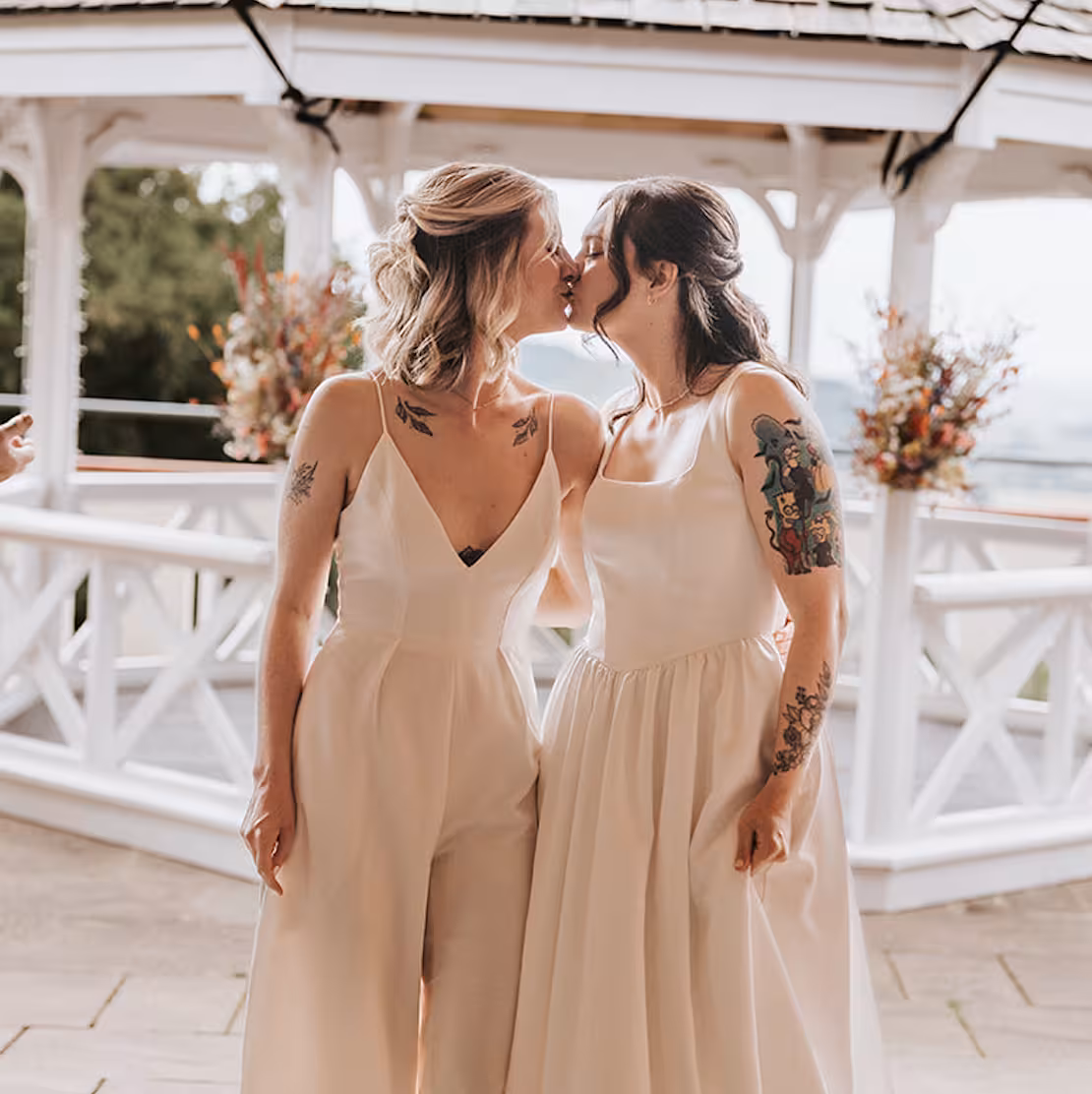 Jodie and Abi share a kiss at their Caer Llan wedding, standing beneath a gazebo decorated with vibrant floral displays, wearing elegant ivory outfits.