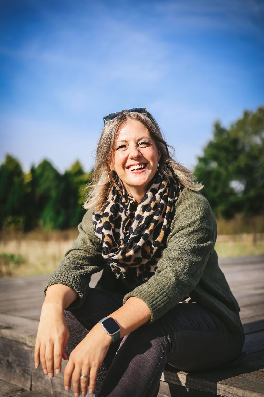 Smiling Kate Rostance sitting outdoors on a sunny day, wearing a green jumper, leopard print scarf, and smartwatch, with trees and blue sky in the background.