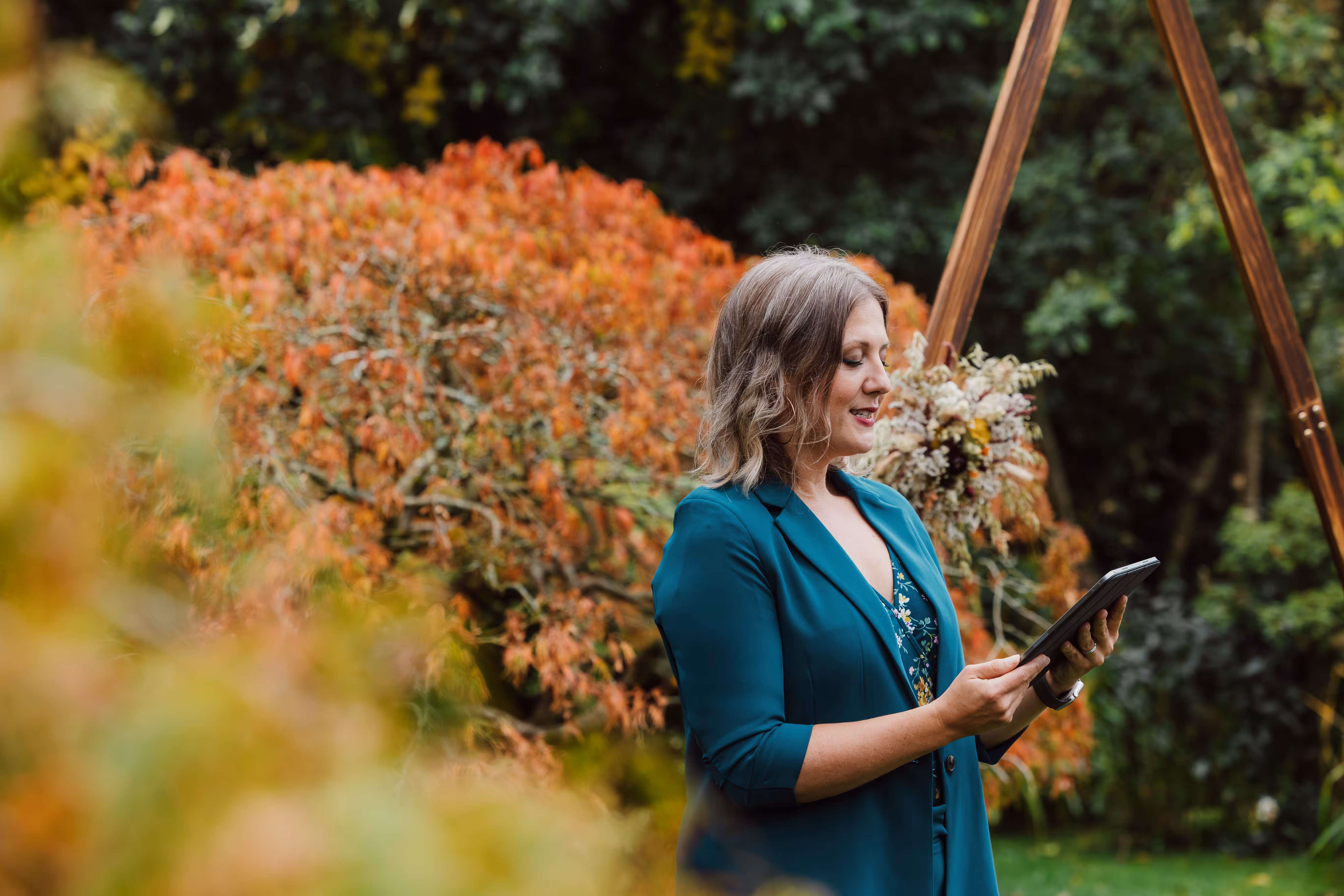 Wedding celebrant leading an autumn ceremony outdoors, standing beside warm orange and gold foliage. She holds a tablet while speaking beneath a wooden arch decorated with dried flowers, perfectly capturing the cosy, colourful atmosphere of an autumn wedding in North Wales.