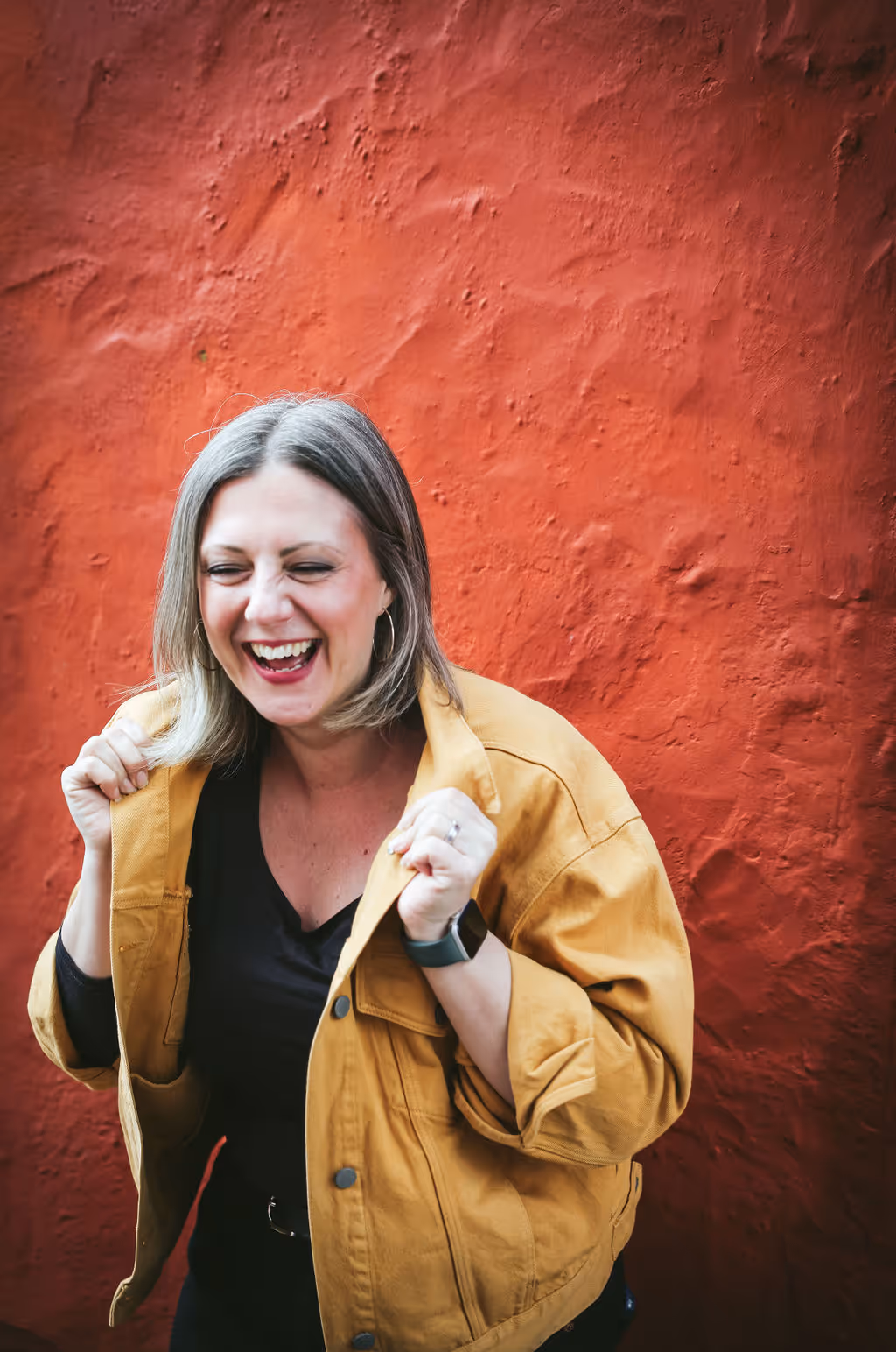 Wedding celebrant Kate Rostance laughing, wearing a mustard yellow jacket against a red wall.