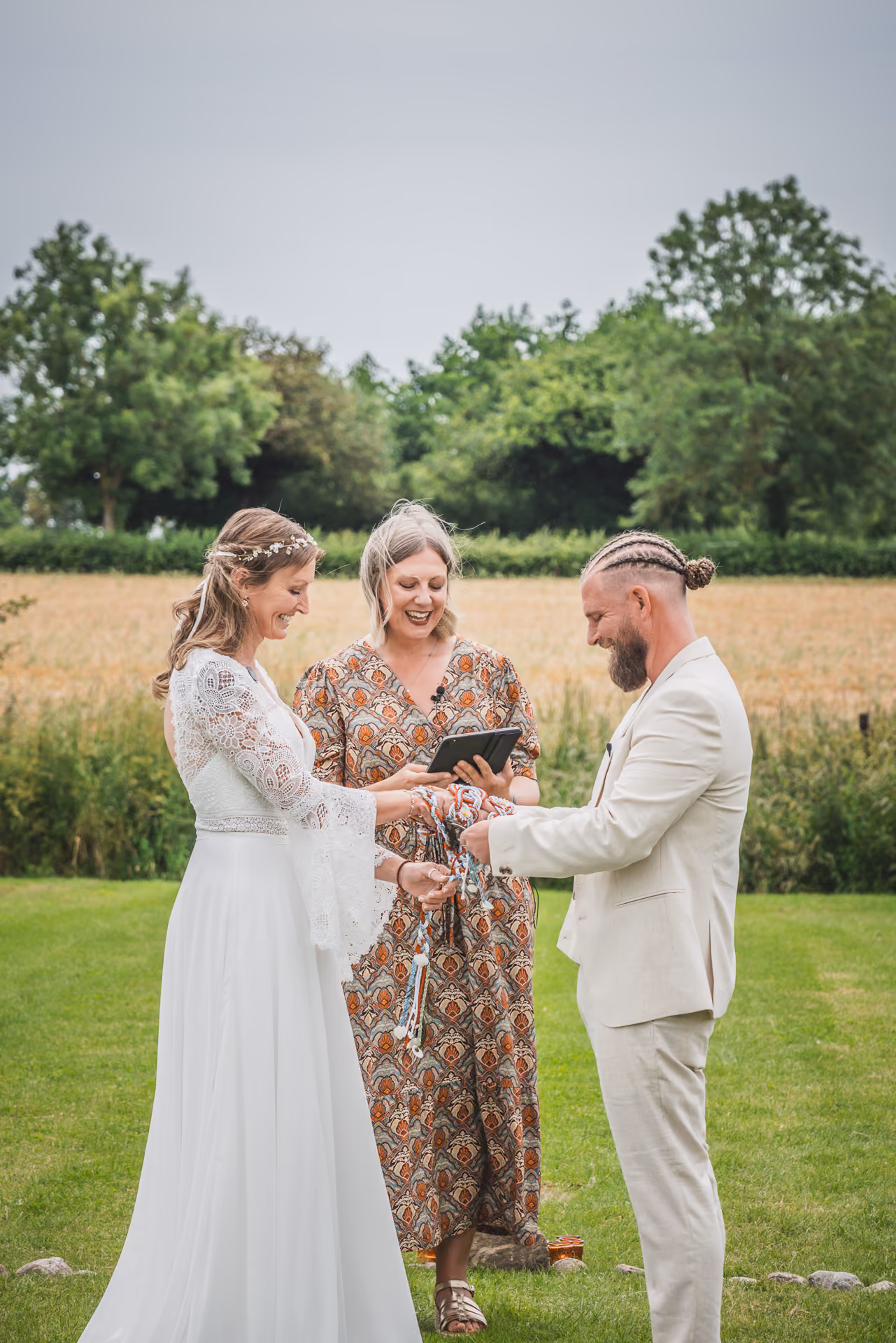 Handfasting ceremony in a Cheshire field, celebrant binding the couple's hands with colourful cords as they laugh together.