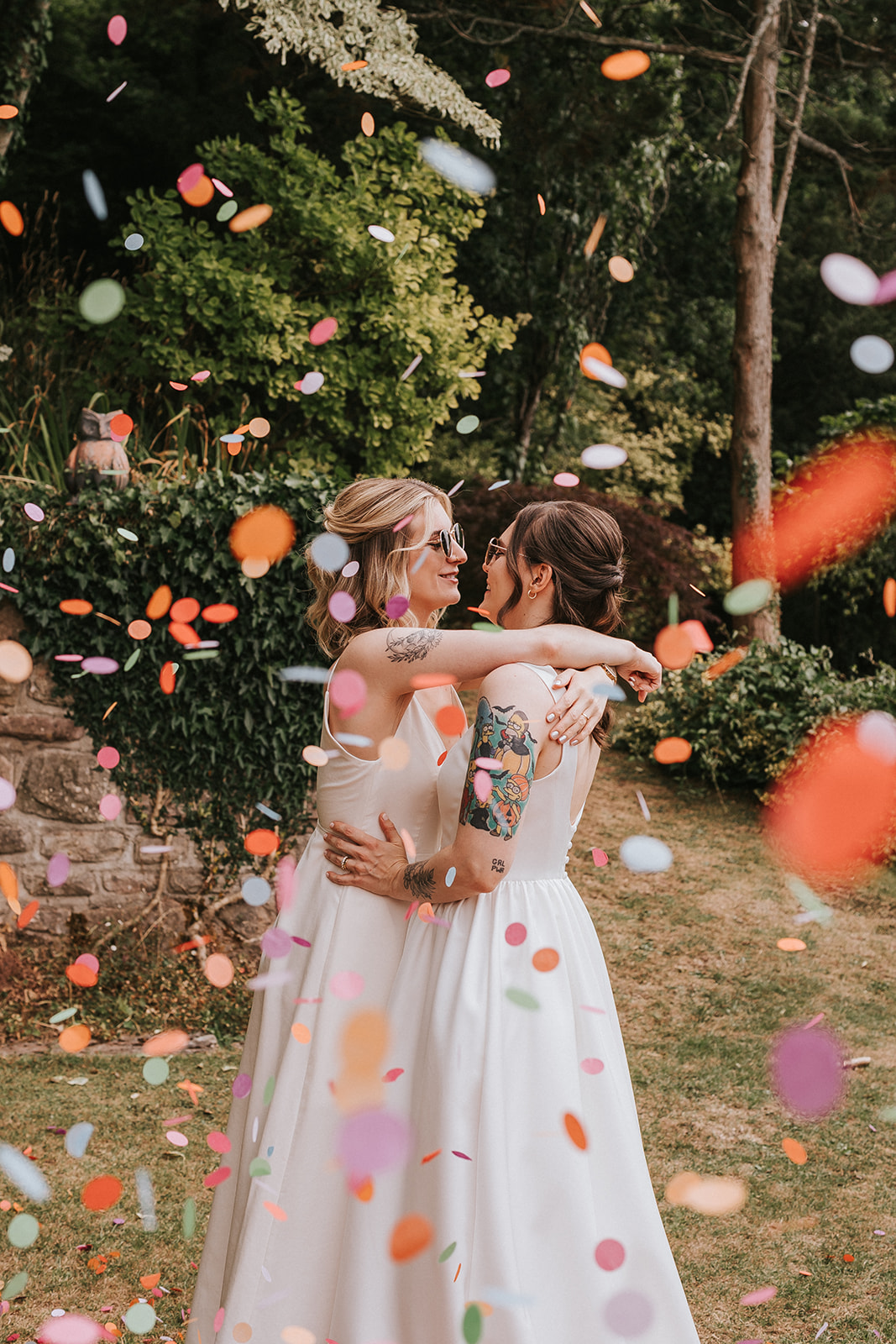 Brides Abi and Jodie hugging during a joyful confetti throw on their wedding day, surrounded by colourful petals in a garden setting.