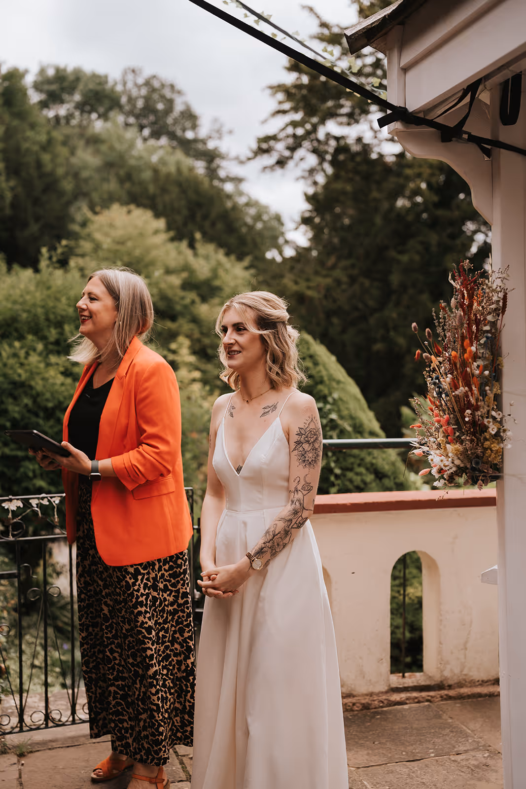 Wedding celebrant Kate Rostance, in an orange jacket, smiling alongside bride Abi during her ceremony at Caer Llan