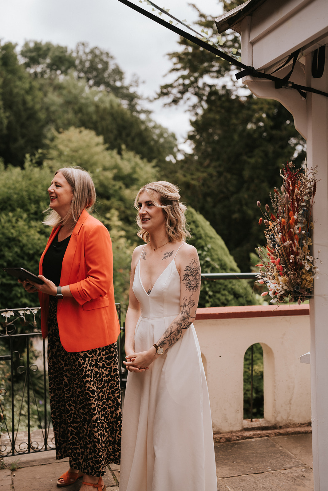 Wedding celebrant Kate Rostance, in an orange jacket, smiling alongside bride Abi during her ceremony at Caer Llan