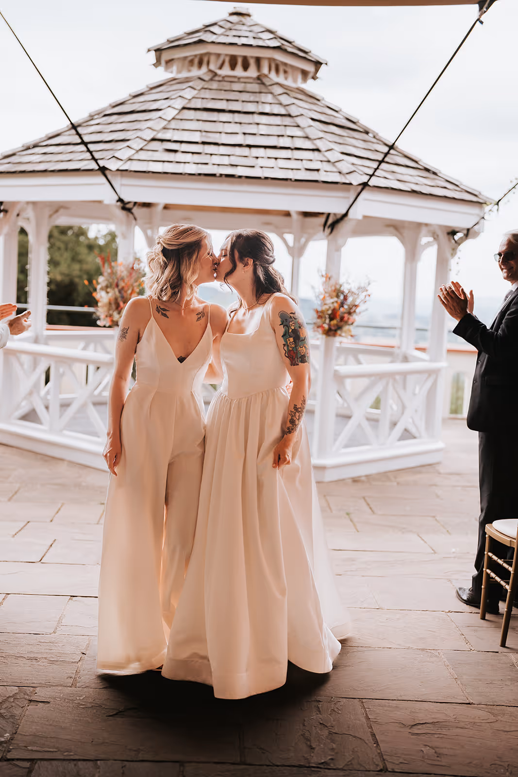 Brides Abi and Jodie sharing a kiss after their wedding ceremony at Caer Llan, standing in front of the white gazebo decorated with flowers as guests applaud.
