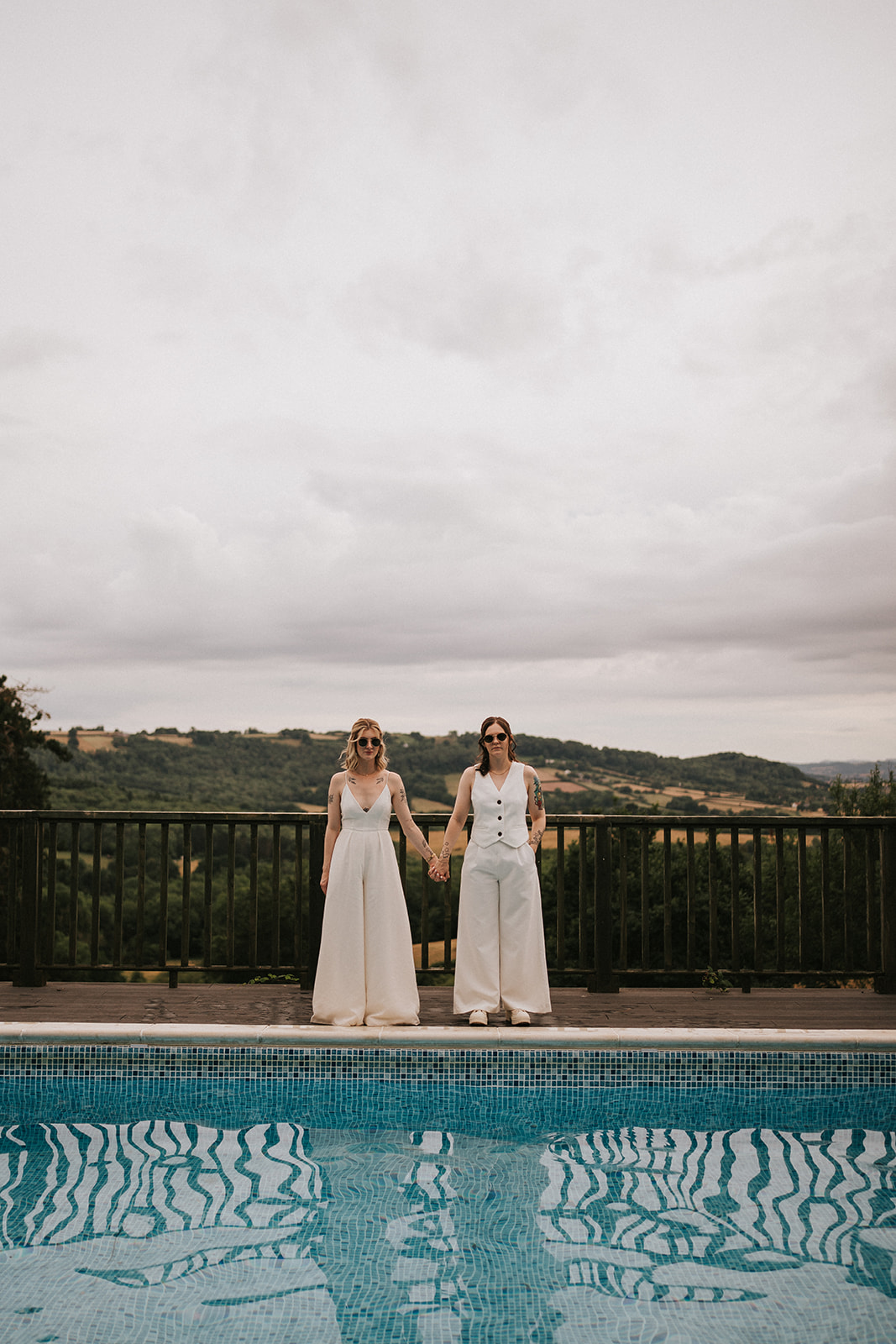 Abi and Jodie holding hands by the pool at Caer Llan on their wedding day, with countryside views in the background.