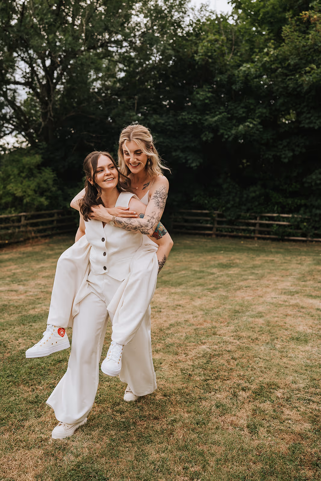Abi giving Jodie a playful piggyback on their wedding day, both laughing in relaxed white outfits during their outdoor celebration at caer llan.