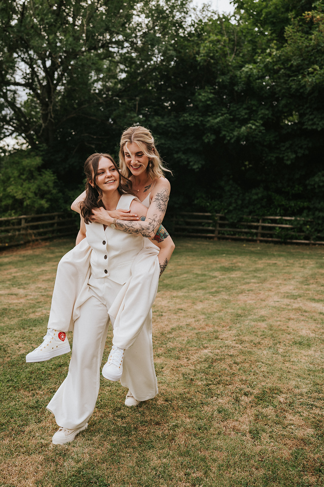Abi giving Jodie a playful piggyback on their wedding day, both laughing in relaxed white outfits during their outdoor celebration at caer llan.
