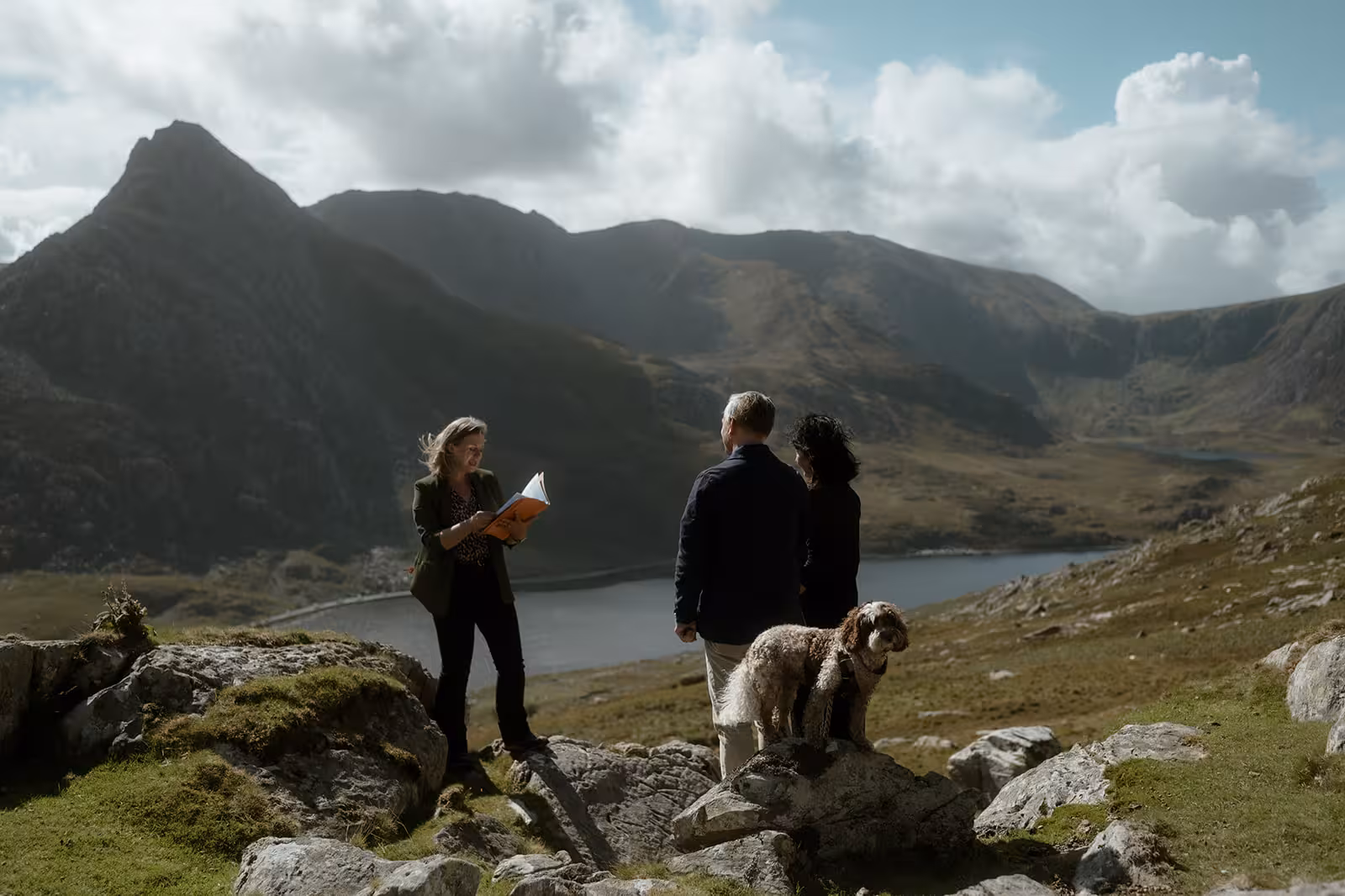 A celebrant leads Gina and Paul’s handfasting ceremony on the mountainside at Pen y Ole Wen, overlooking the lake, with their dog Coco beside them under dramatic skies.