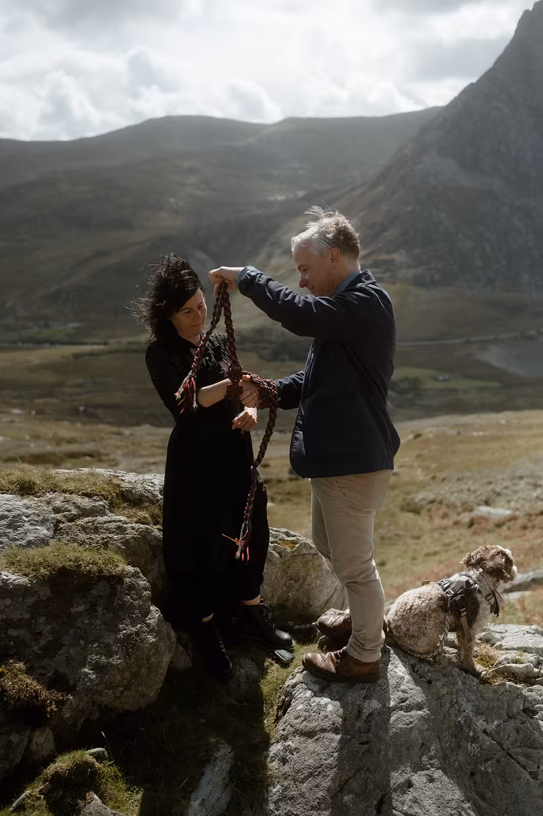 Gina and Paul perform their handfasting ritual on Pen y Ole Wen, wrapping red cords around their joined hands as their dog Coco sits beside them on the mountain rocks.