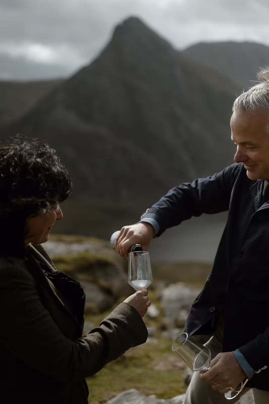 Paul pours fizz into Gina’s glass as they toast their handfasting ceremony, with the dramatic mountain peak of Pen y Ole Wen rising behind them in Eryri.
