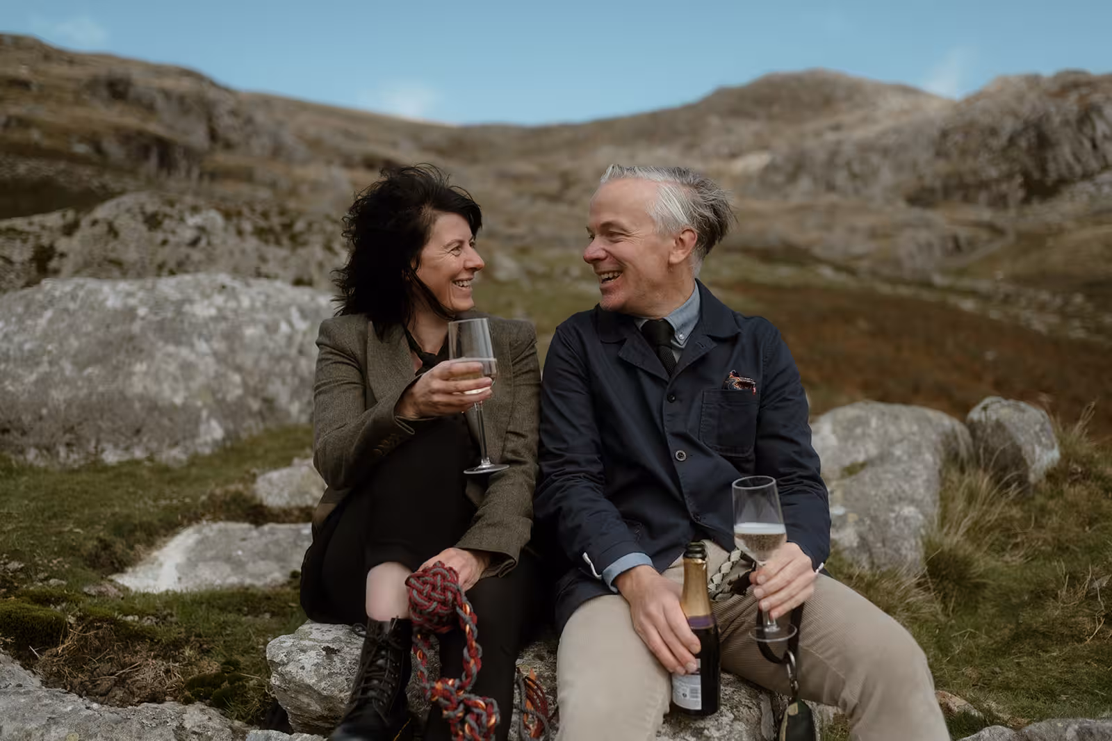 Gina and Paul sit on a rock with glasses of fizz, laughing together after their handfasting ceremony on Pen y Ole Wen in Eryri, surrounded by rugged mountain scenery.