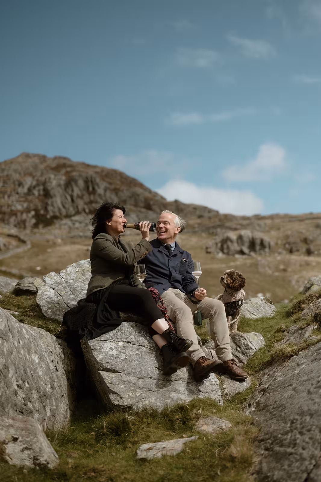 Gina and Paul share a drink and laugh together while sitting on a rock with their dog Coco beside them, celebrating their wild handfasting ceremony on Pen y Ole Wen in Eryri.