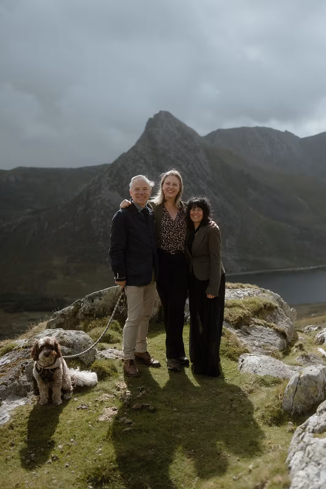 Gina and Paul stand arm in arm with their celebrant on Pen y Ole Wen, smiling with their dog Coco by their side and the dramatic Eryri peaks behind them.