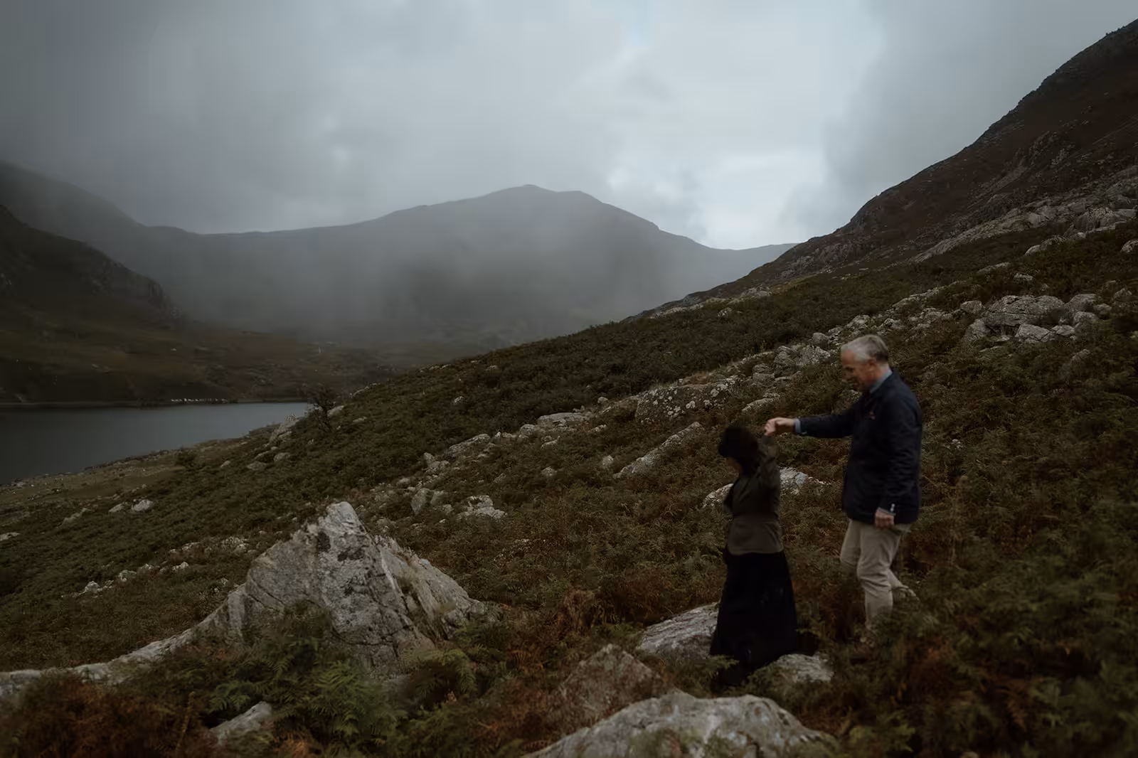 Paul helps Gina carefully navigate the rocky hillside above Llyn Ogwen in Eryri, walking together through the mist after their wild mountain handfasting ceremony.