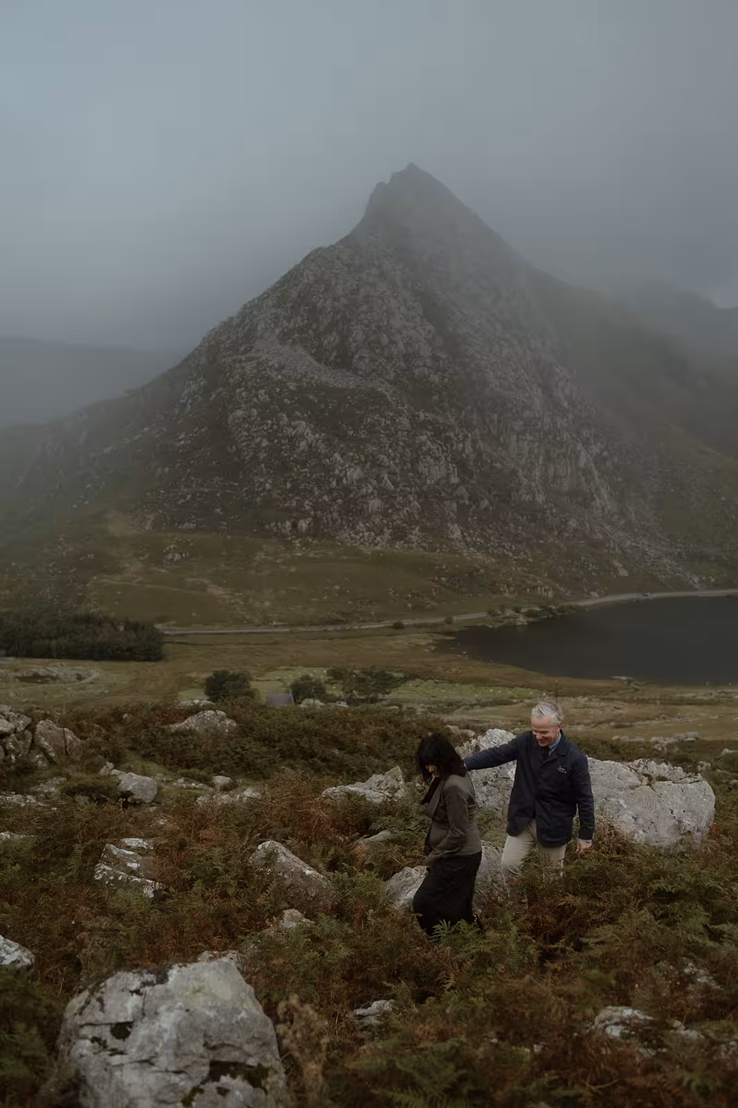 Gina and Paul climb the misty hillside of Pen y Ole Wen after their handfasting ceremony, with the mountain rising steeply behind them and the lake below.