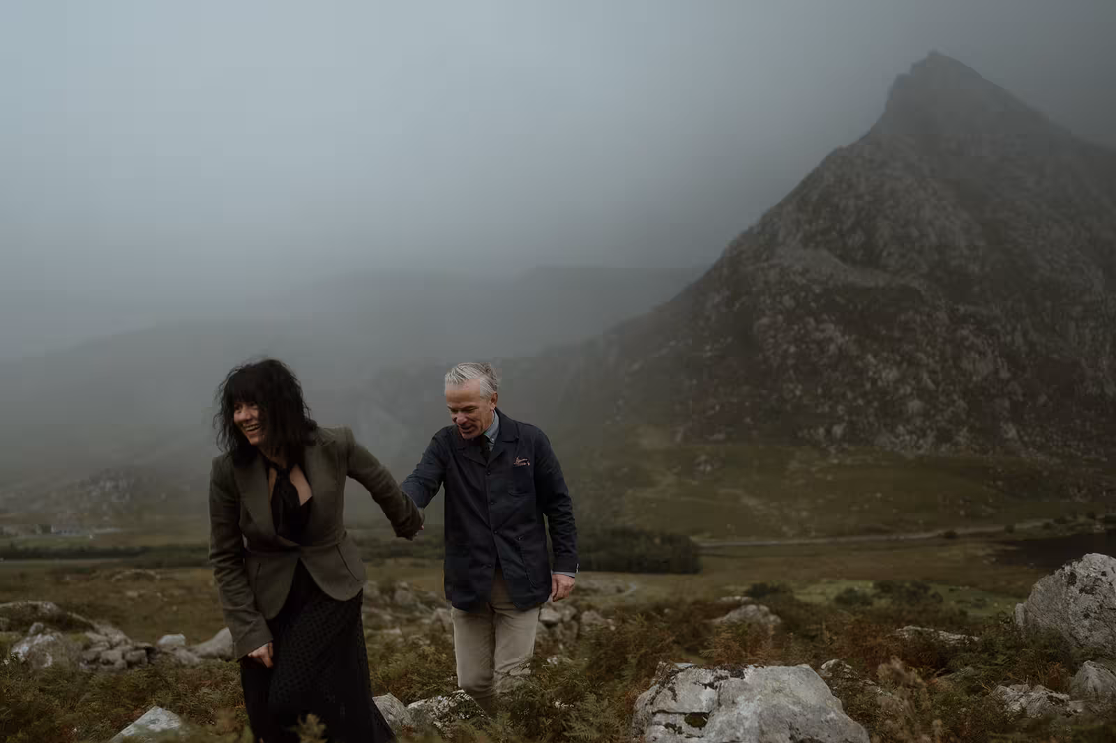 Gina and Paul laugh and hold hands as they hike up the rocky slopes of Pen y Ole Wen, the misty peak towering behind them in Eryri National Park.