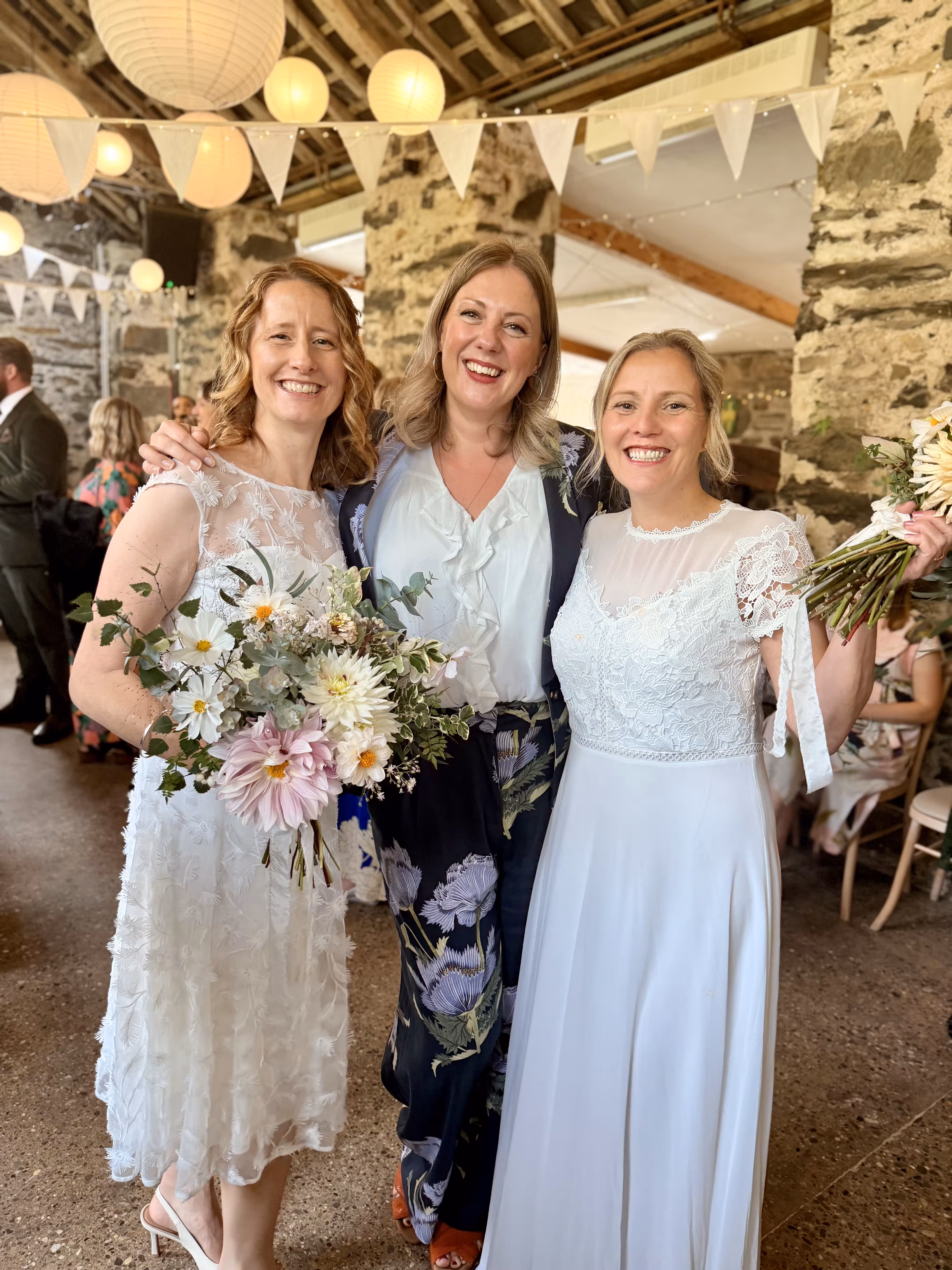 Celebrant Kate Rostance with newlyweds Clair and Janine after their humanist wedding ceremony at Llyn Gwynant Barns in North Wales, holding bouquets of fresh wildflowers.