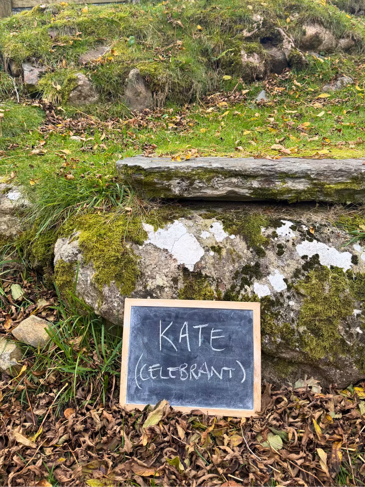 Chalkboard sign reading ‘Kate (Celebrant)’ placed outdoors on mossy stone steps at Llyn Gwynant Barns in Gwynedd.