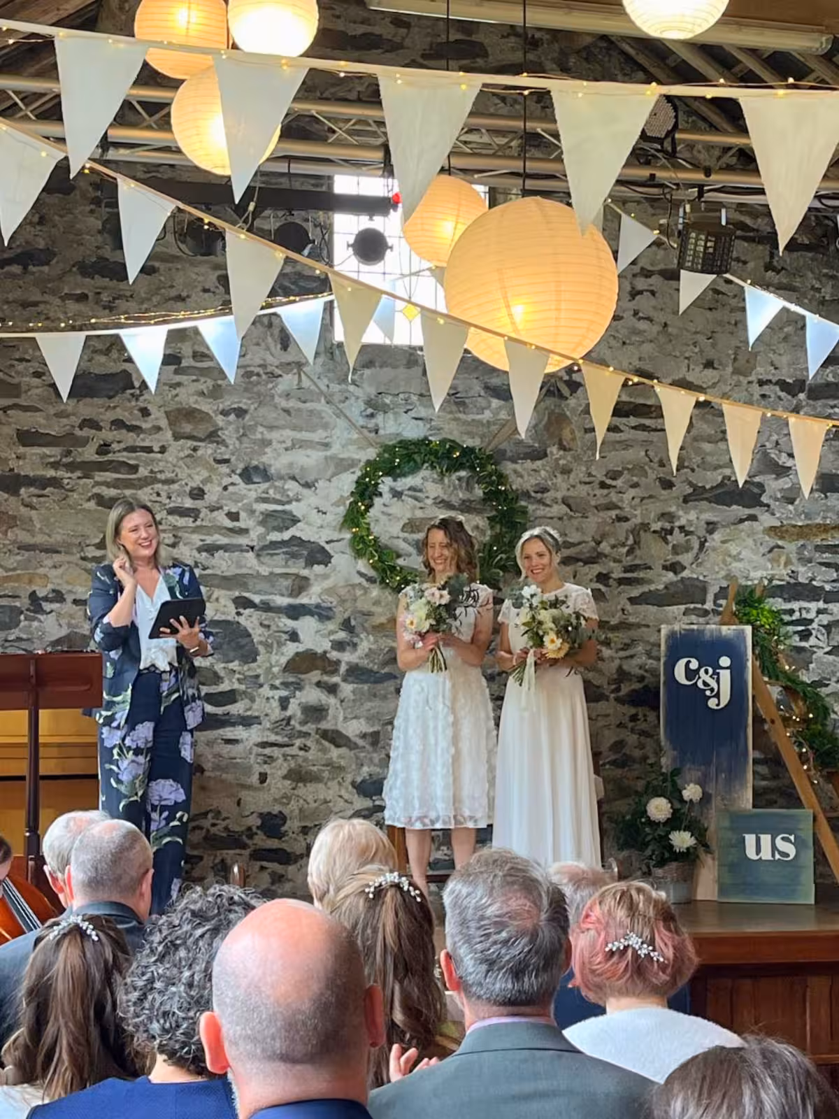 Wedding celebrant leading Clair and Janine’s same-sex ceremony at Llyn Gwynant Barns in Gwynedd, surrounded by rustic stone walls, bunting and lanterns during their autumn barn wedding.