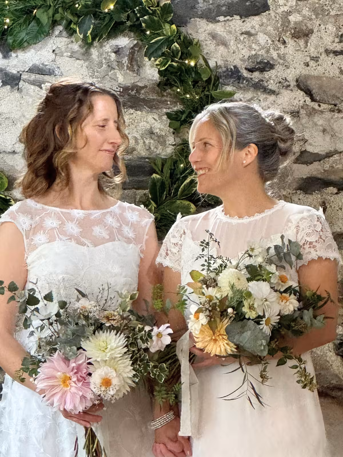 Brides Clair and Janine standing hand in hand with wildflower bouquets during their humanist wedding ceremony at Llyn Gwynant Barns in Gwynedd.