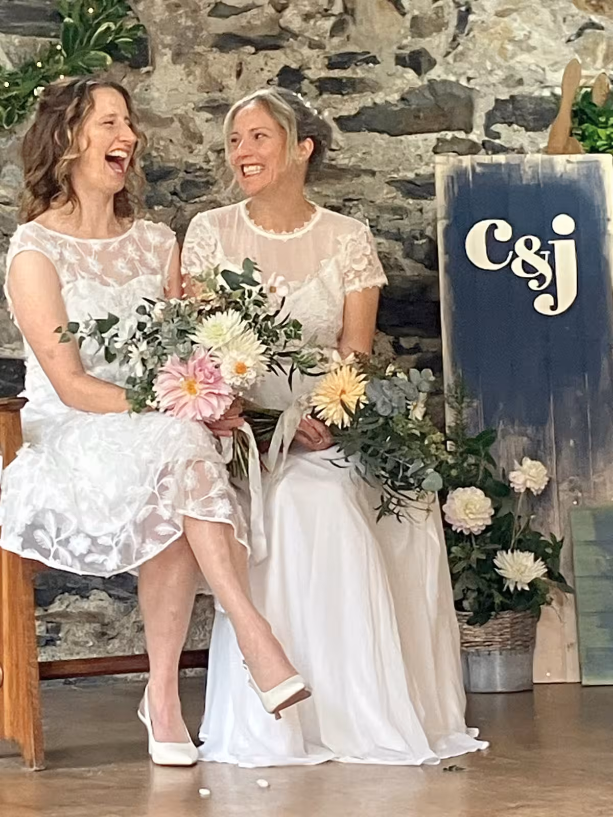 Brides Clair and Janine laughing together during their same-sex wedding ceremony at Llyn Gwynant Barns, with flowers and rustic decor behind them