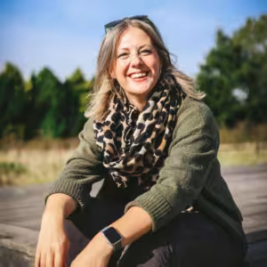 Kate Rostance, a smiling humanist celebrant based in North Wales, sitting outdoors on a sunny day wearing a green jumper and leopard-print scarf, used as an author profile photo for her wedding and funeral celebrant blog.