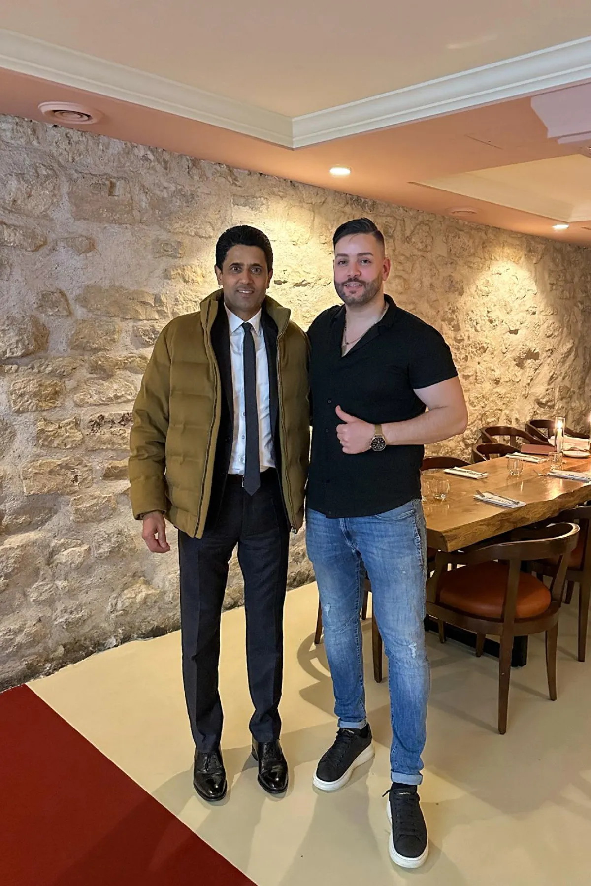 Two men standing side by side in a restaurant with stone walls, one in a brown jacket and formal attire, the other in a black shirt and jeans giving a thumbs up.