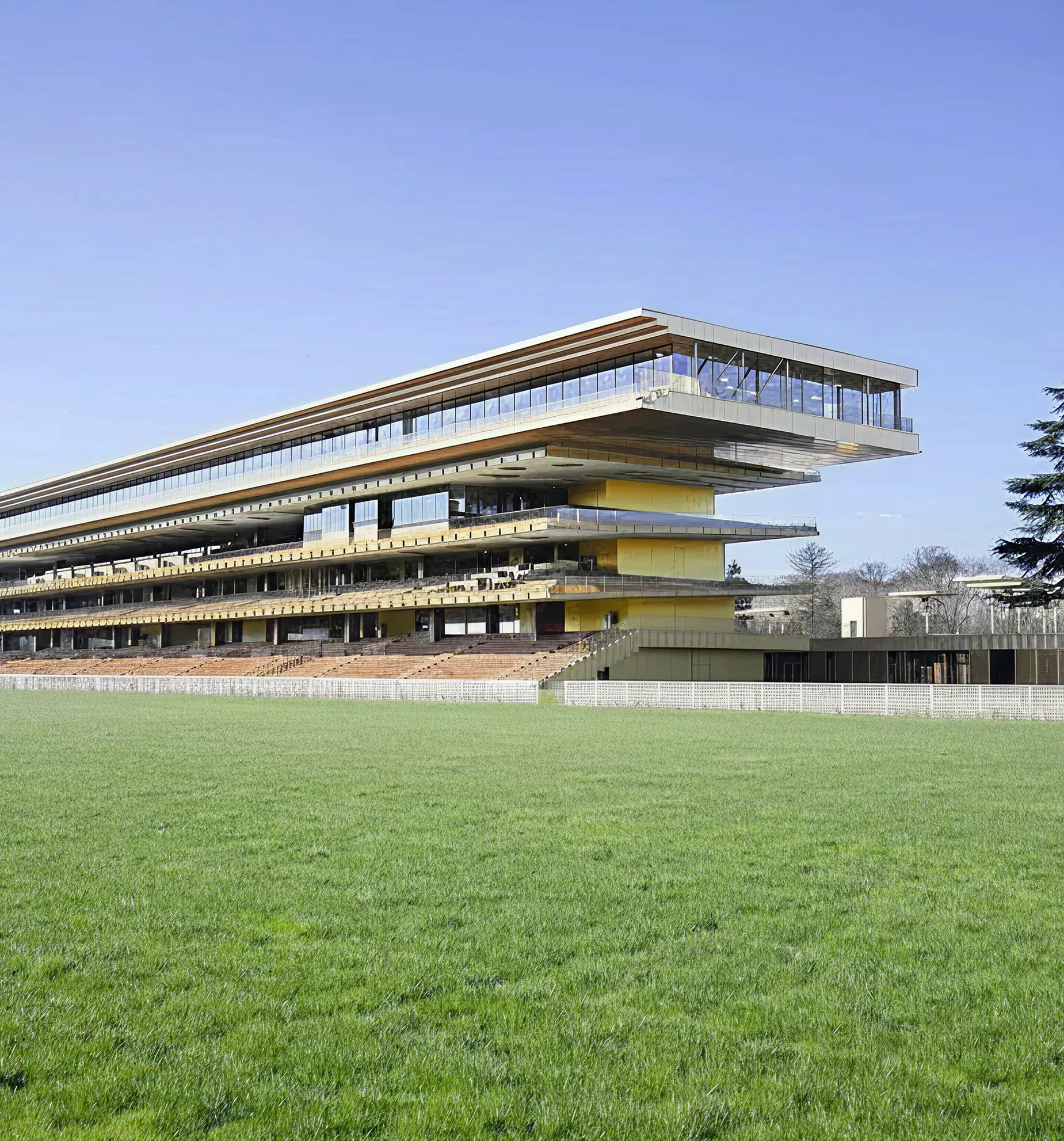 Modern multi-level building with extended cantilevered glass section, yellow walls, and wide balconies overlooking a large green grass field.