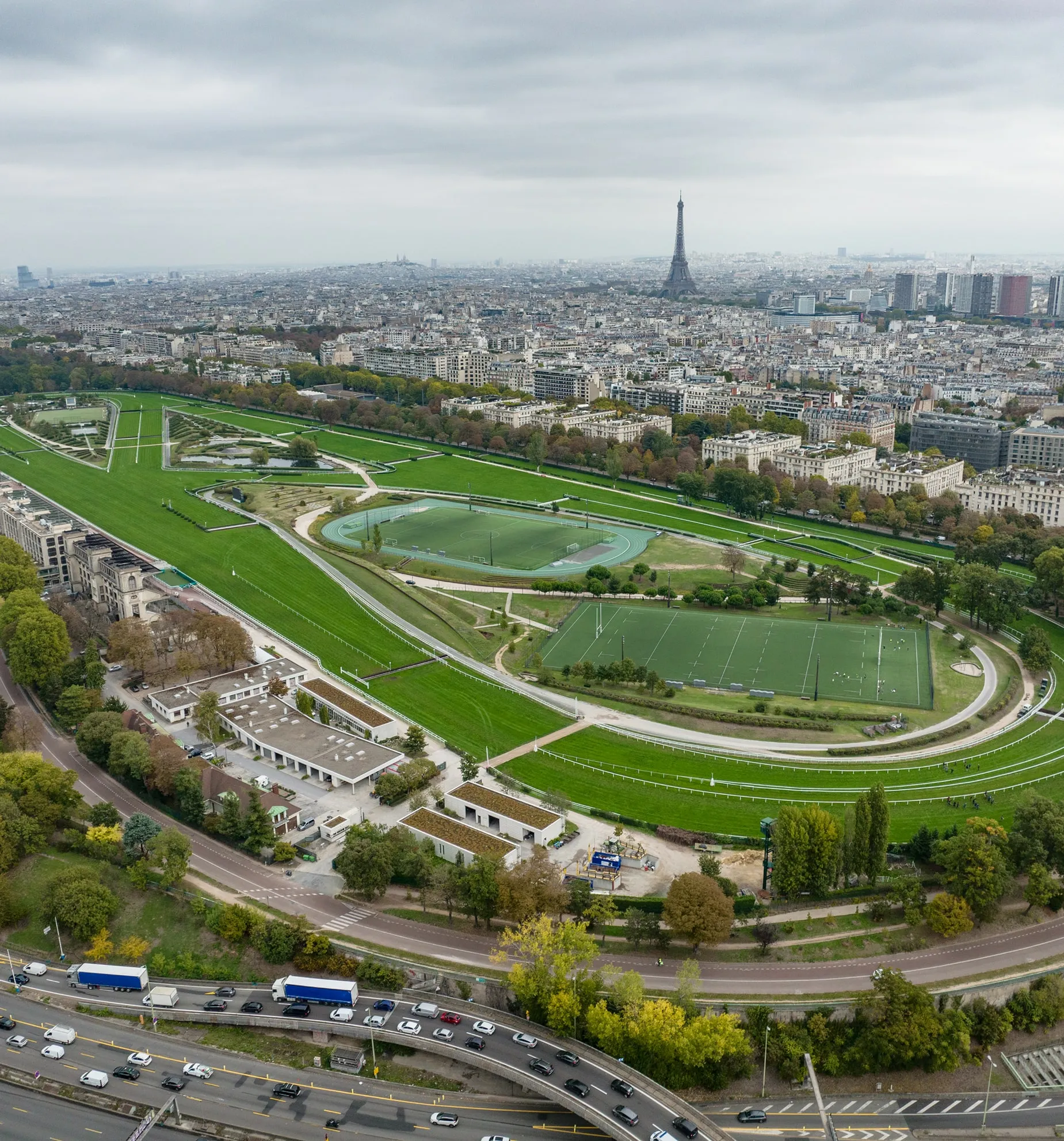 Aerial view of a large green sports complex with fields and tracks, with the Eiffel Tower and Paris cityscape in the background under a cloudy sky.