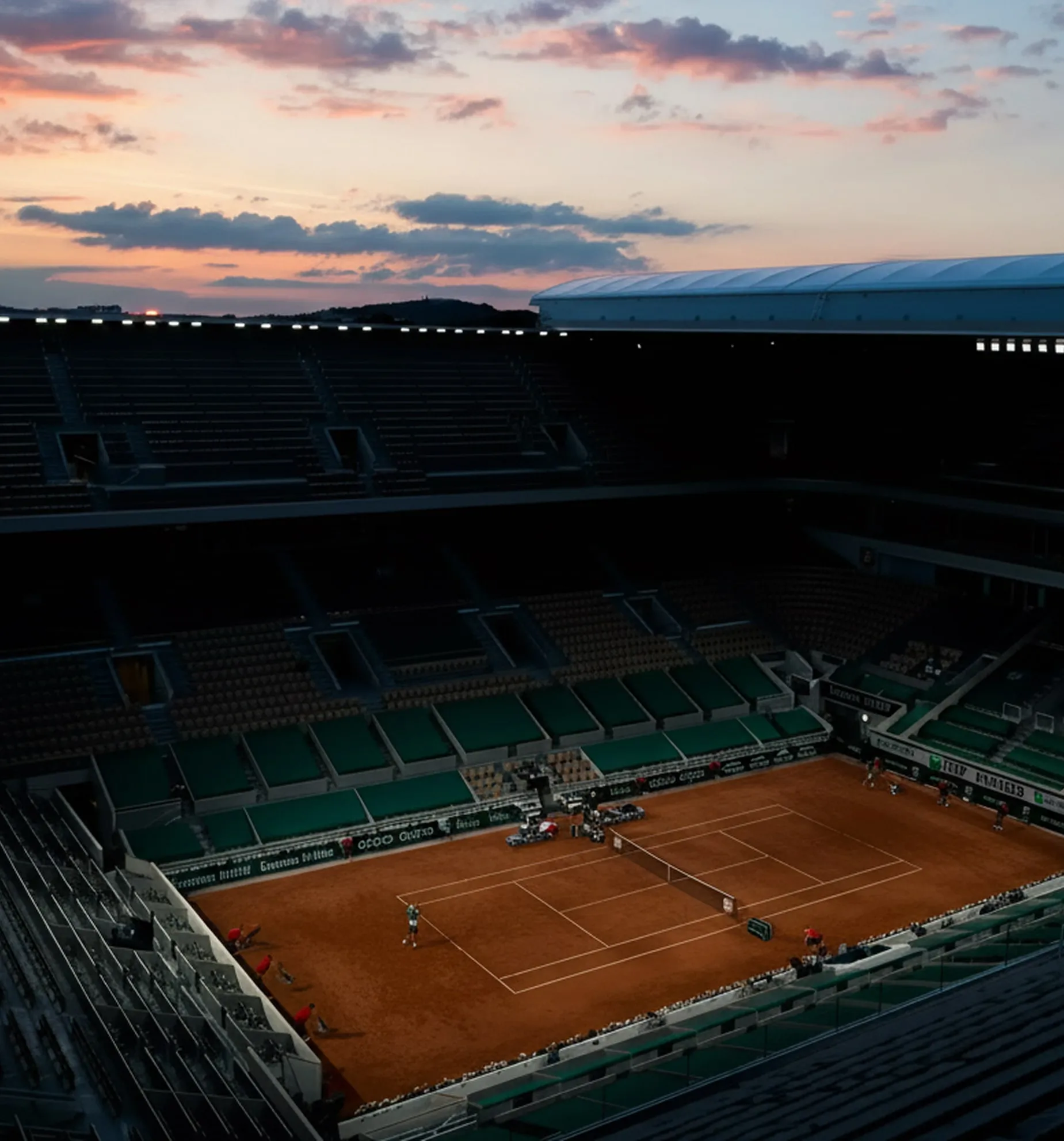 Empty tennis stadium with clay court under a colorful sunset sky.