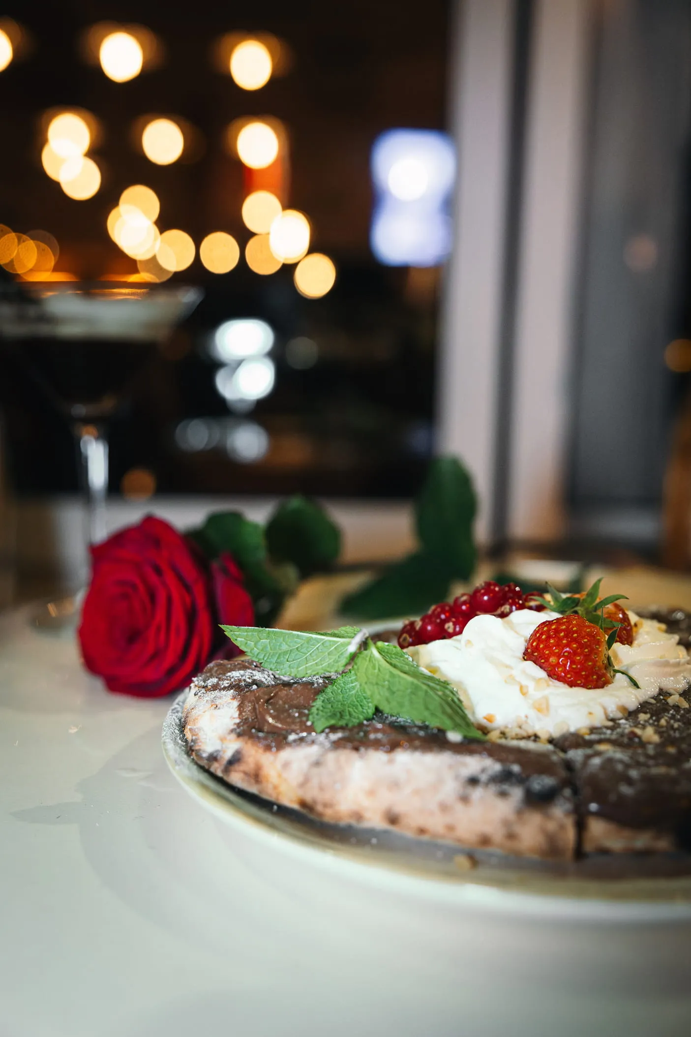 Close-up of a dessert pizza topped with whipped cream, strawberries, red currants, and mint leaves, with a red rose and a cocktail glass in the blurred background.