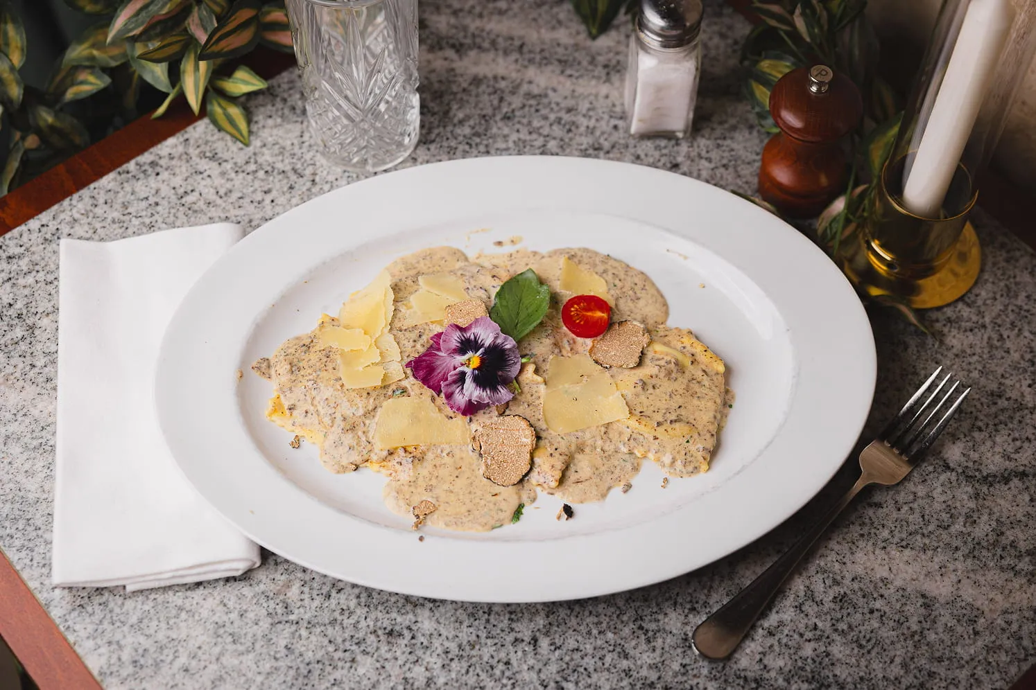 Plate of ravioli covered in creamy truffle sauce, garnished with shaved cheese, truffle slices, a purple edible flower, a green leaf, and half a cherry tomato on a granite table with a fork and folded napkin.