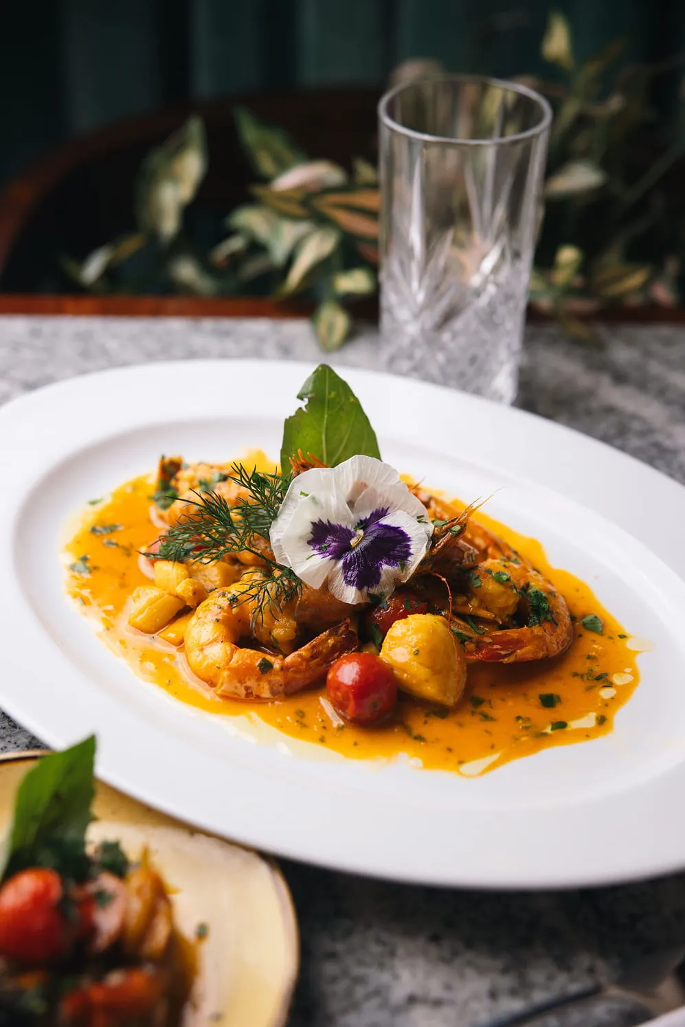 Plate of shrimp in orange sauce garnished with a white and purple edible flower, herbs, and cherry tomatoes.