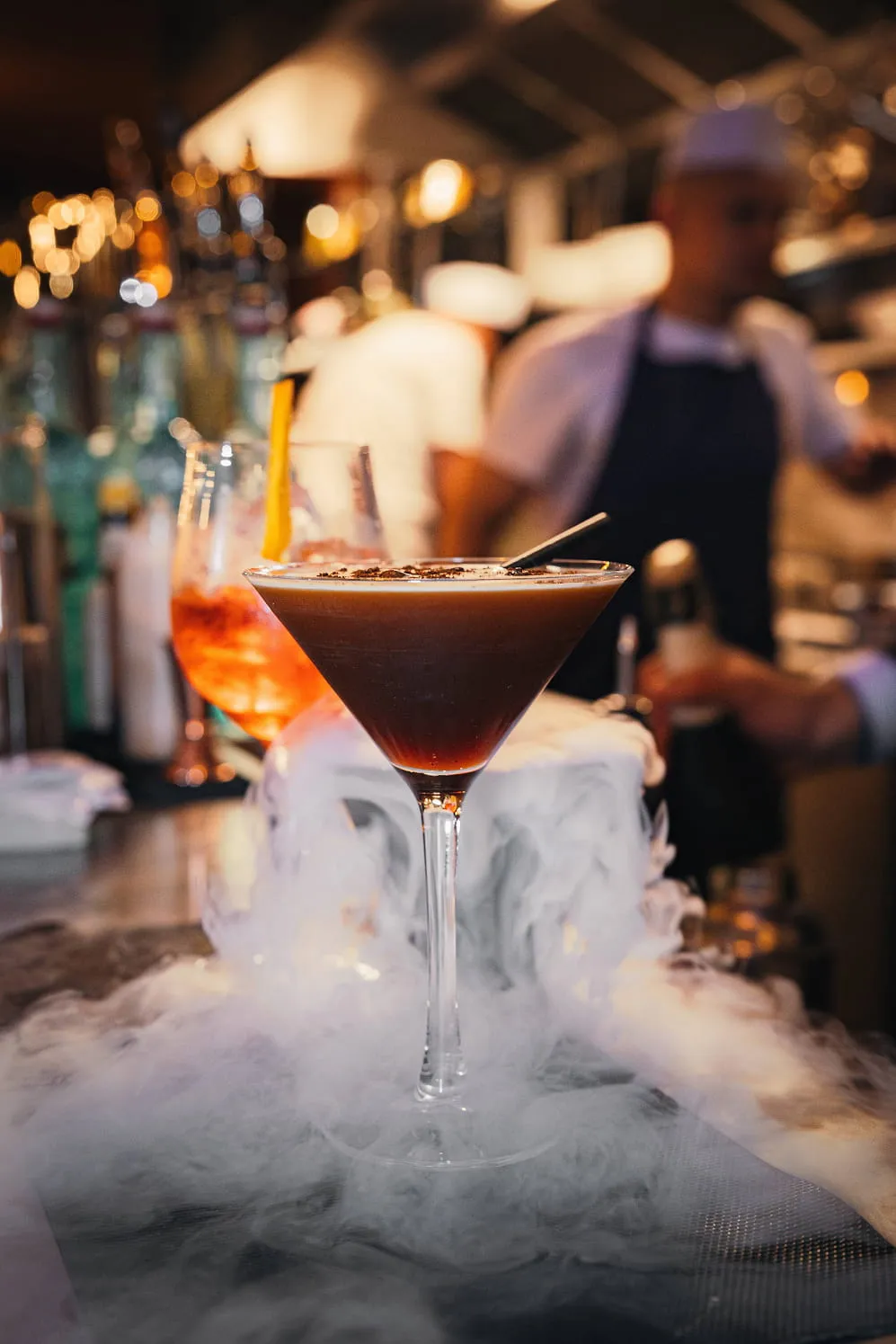 Espresso martini cocktail in a martini glass surrounded by mist or smoke on a bar counter with blurred bartender and bottles in the background.