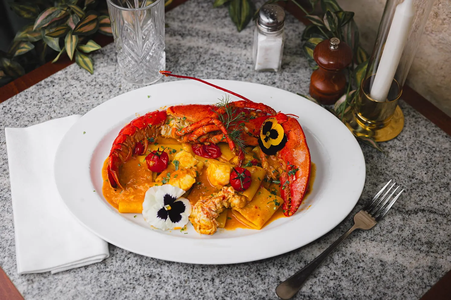 Plate of paccheri pasta with lobster claws, cherry tomatoes, herbs, and edible flowers on a granite table with a fork and napkin.