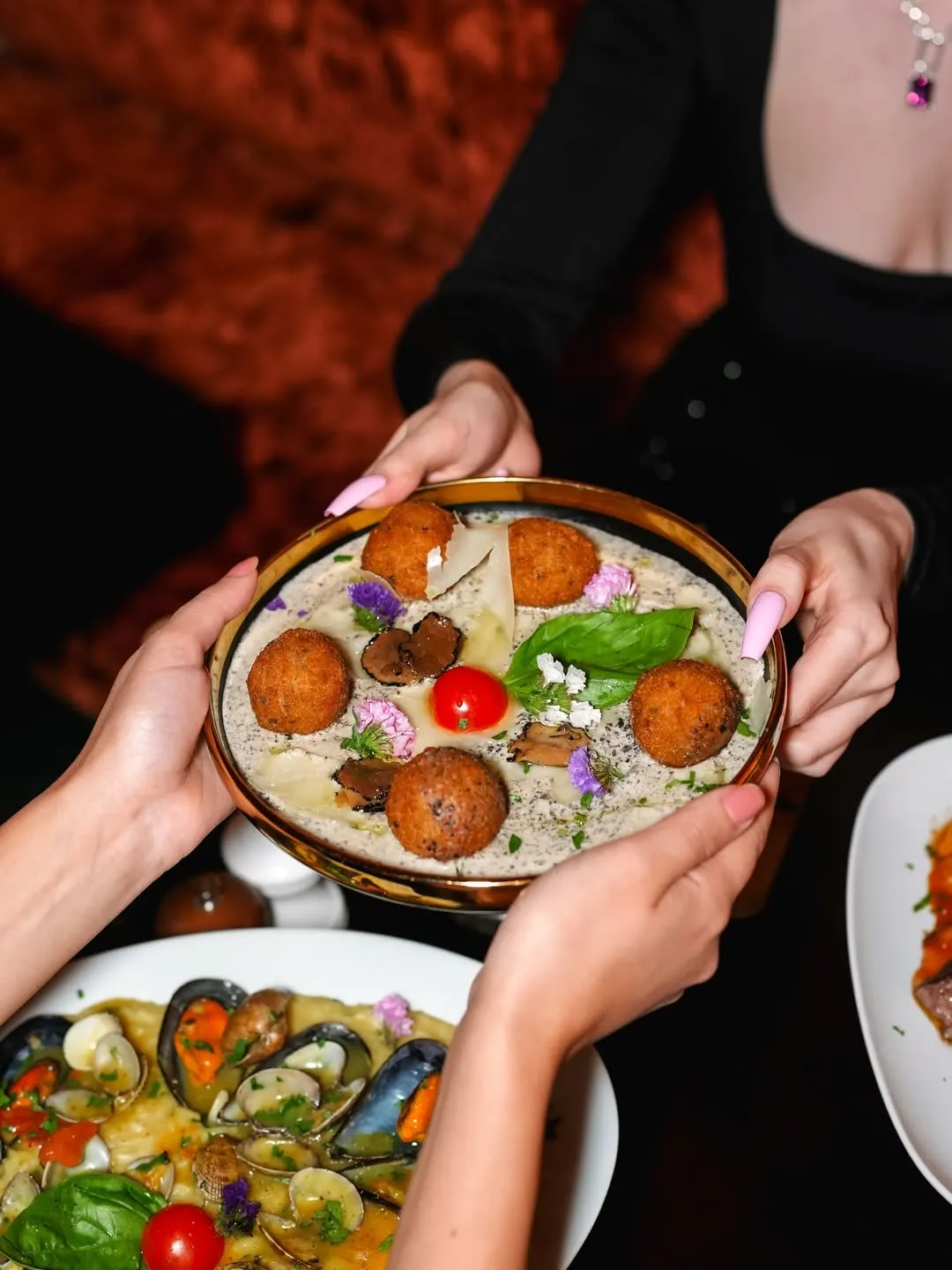 Two people exchanging a plate with five fried croquettes on creamy sauce garnished with tomato, basil, mushrooms, and edible flowers.