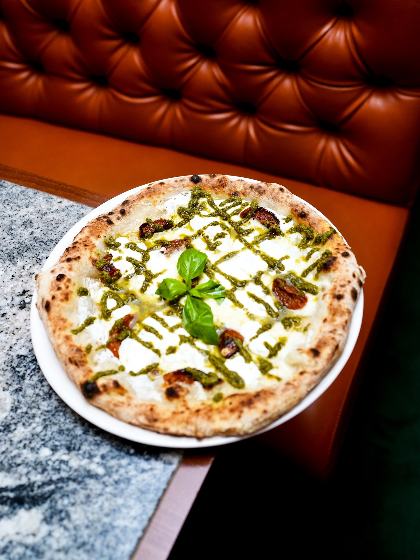 Rustic pizza topped with melted white cheese, pesto drizzle, sun-dried tomatoes, and fresh basil leaves on a white plate.