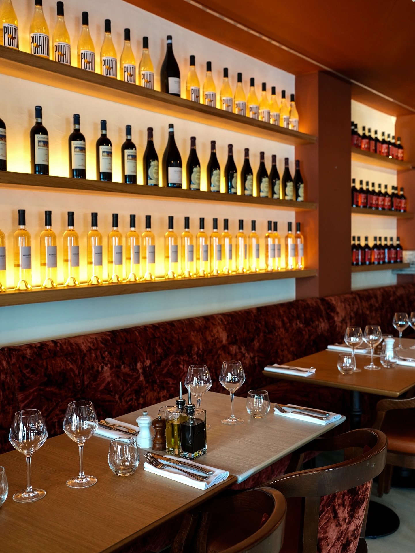 Cozy restaurant interior with wooden tables, velvet seating, wine glasses, and shelves of illuminated wine bottles on the wall.