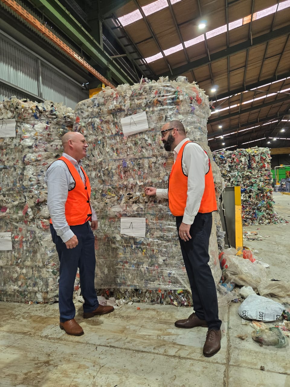 Two men in orange safety vests talking in a recycling warehouse near large bales of compressed plastic waste.