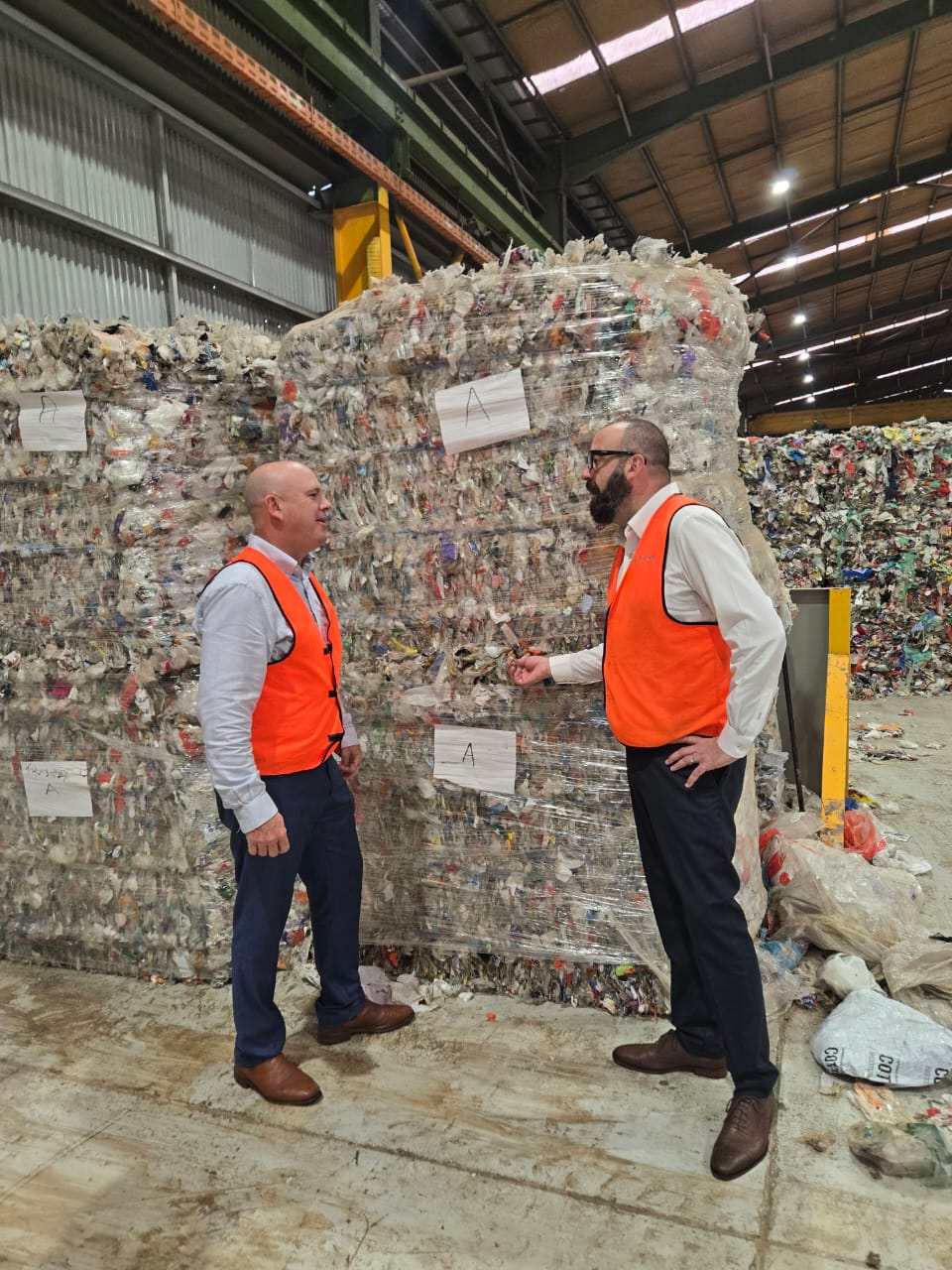 Two men in orange safety vests standing in a recycling facility in front of large packed plastic waste bales.