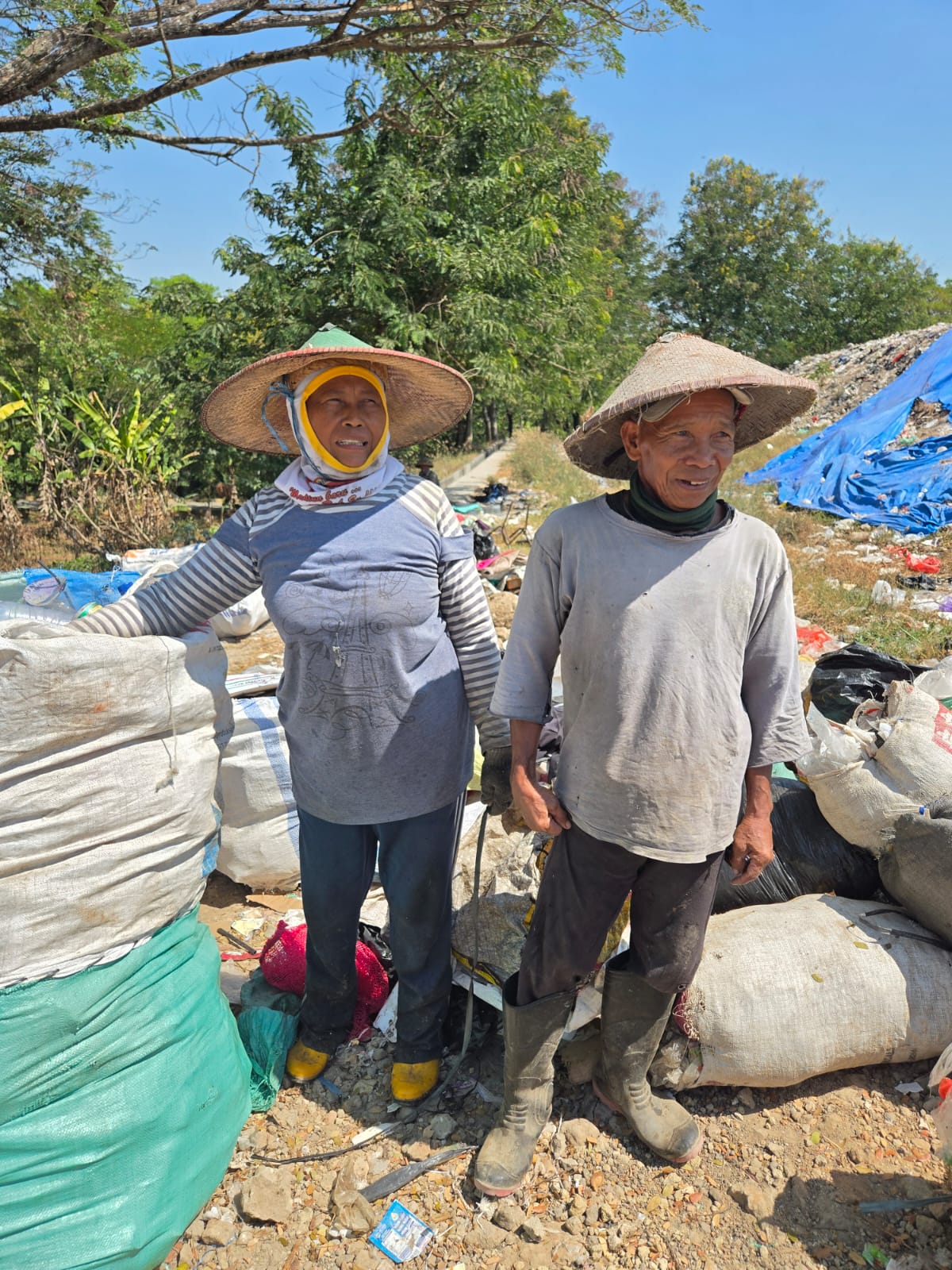 Two older workers wearing wide hats standing outdoors near bags and debris under clear blue sky.