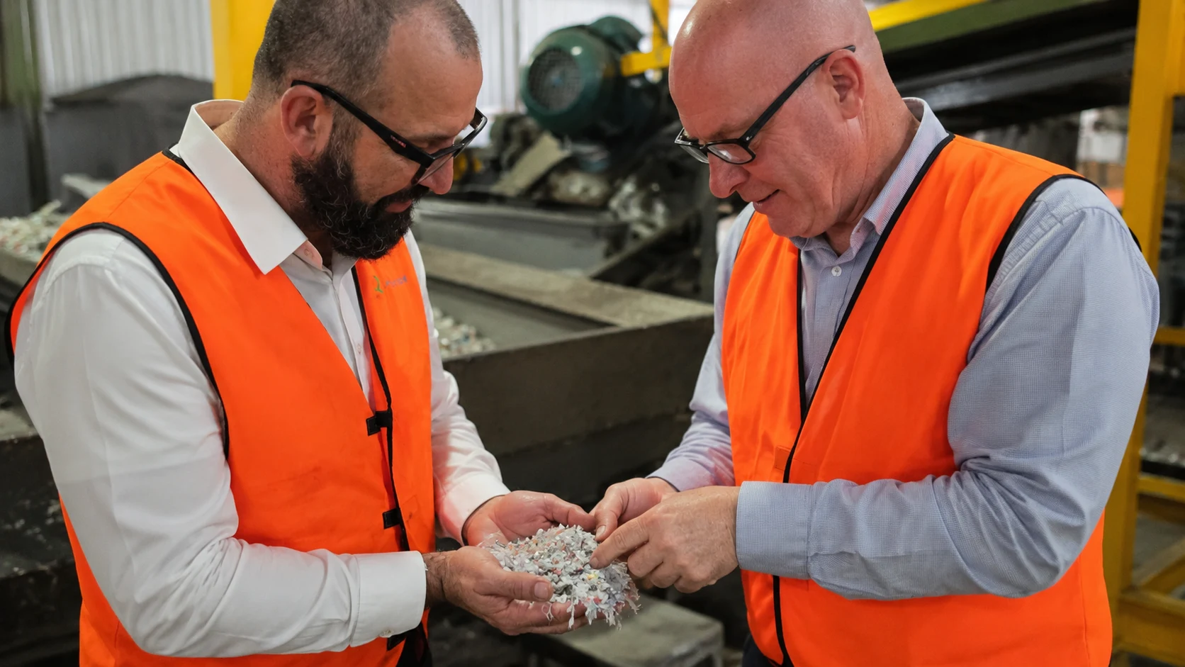 Two men in orange safety vests inspecting shredded plastic pieces in a factory setting.
