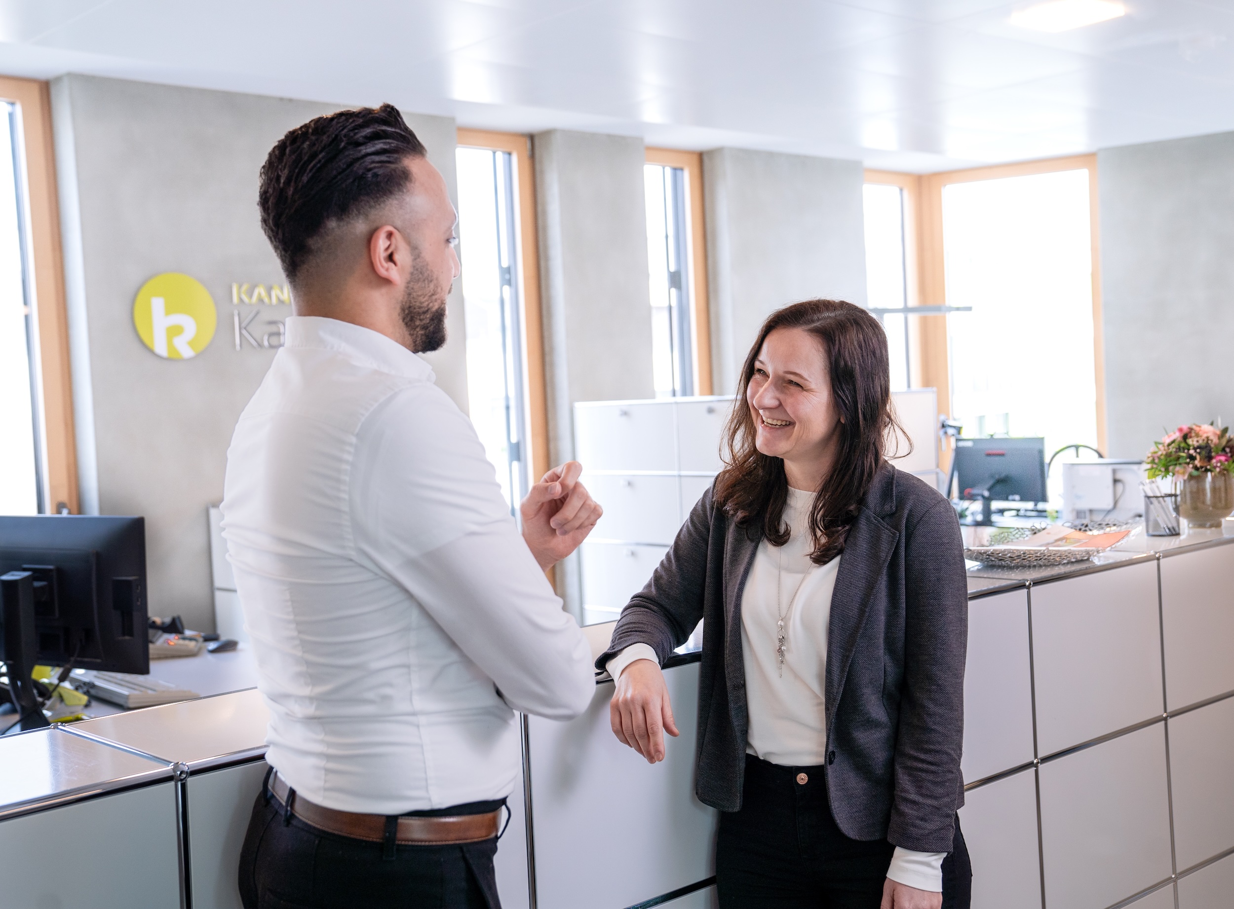Zwei Personen sprechen im Büro, Mann gestikuliert, Frau hört zu.