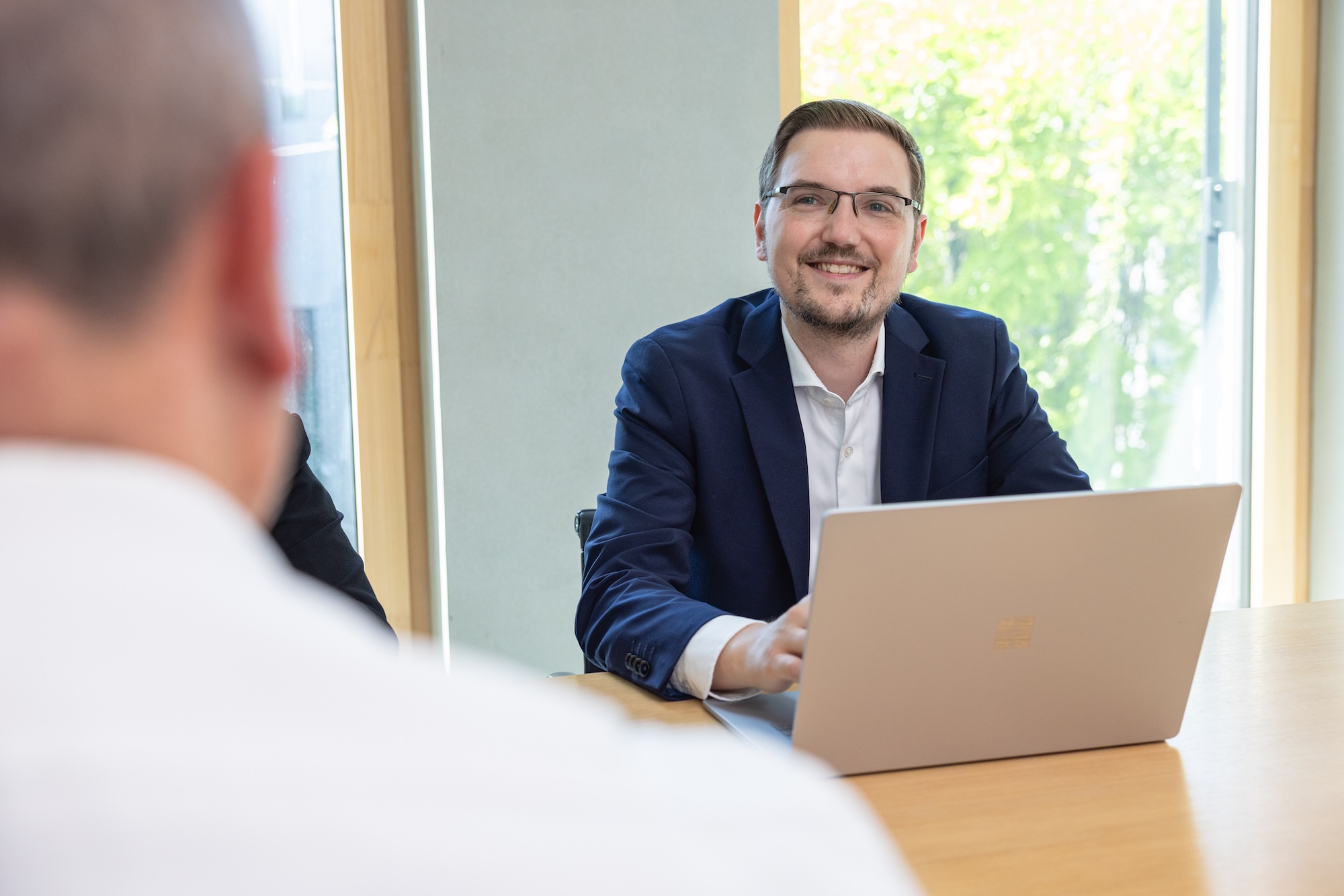 Meeting-Situation im Büro, Person sitzt am Tisch mit Laptop und lächelt.