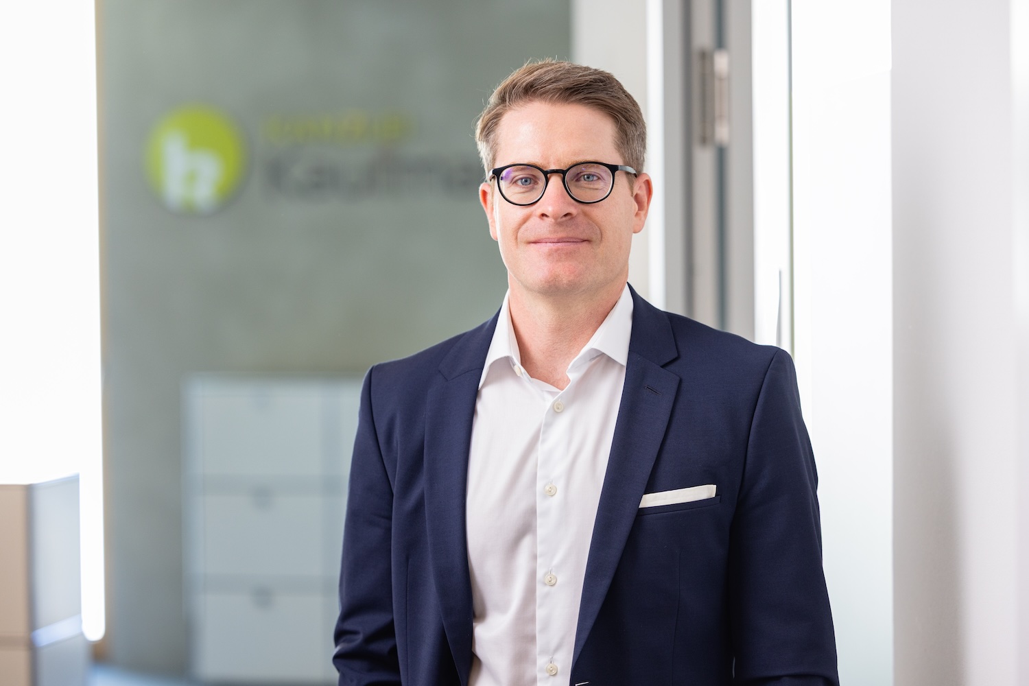 Porträt eines Mannes im Anzug im Büro vor Glaswand mit Logo.